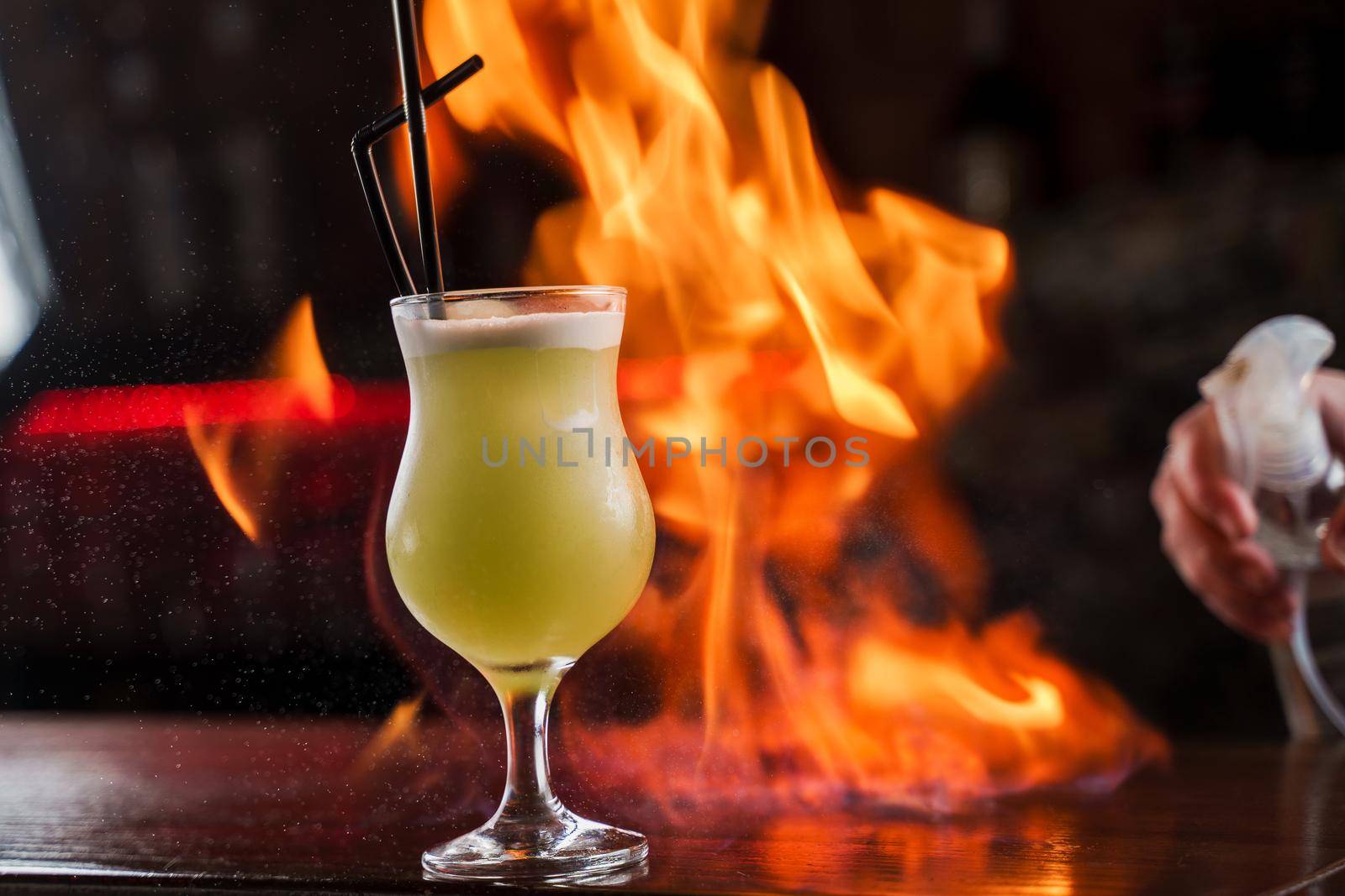 Bartender sprinkles on illuminated glass with bright green cold cocktail on bar counter and makes fire flame over it.
