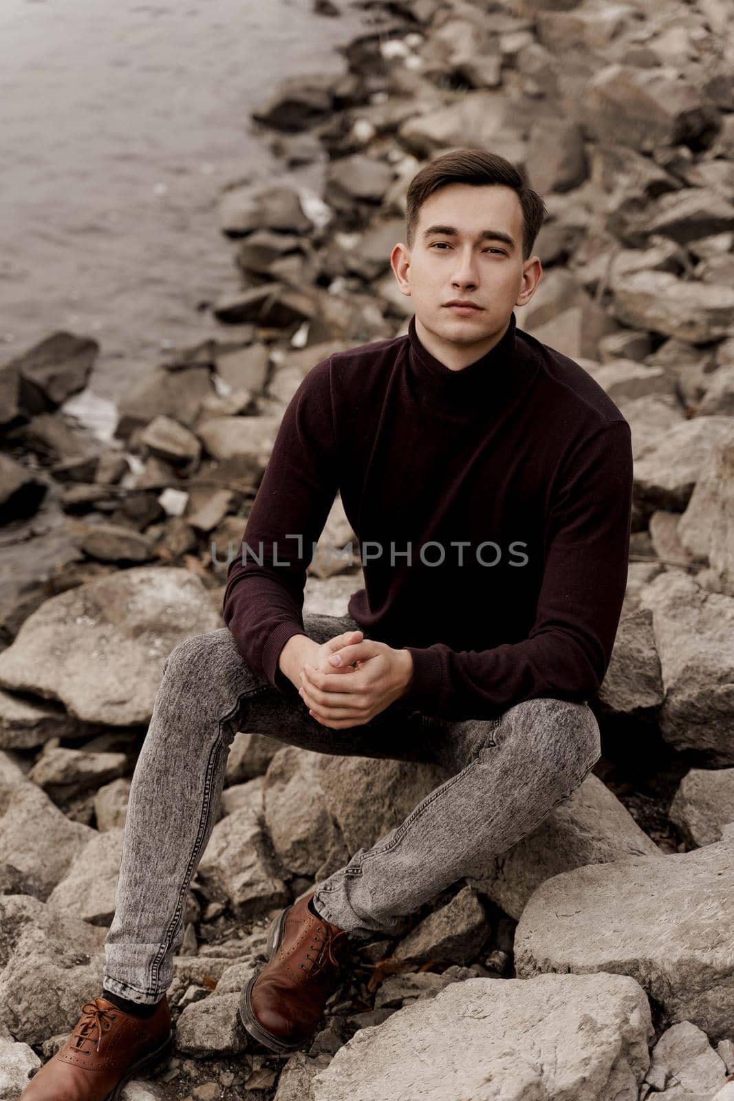 Handsome man seats on the rocks near lake. Professional male model weared formal pullower