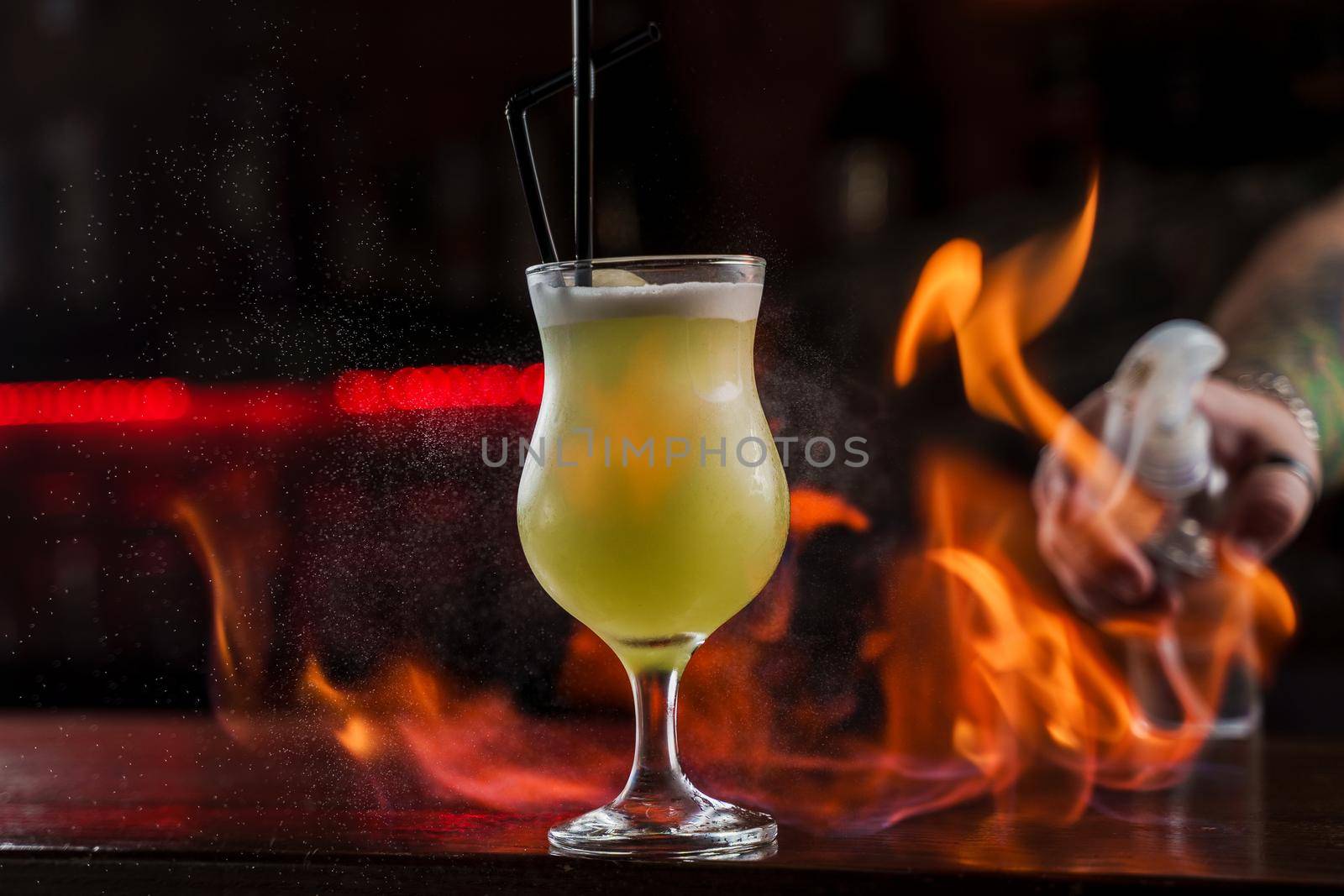 Bartender sprinkles on illuminated glass with bright green cold cocktail on bar counter and makes fire flame over it.