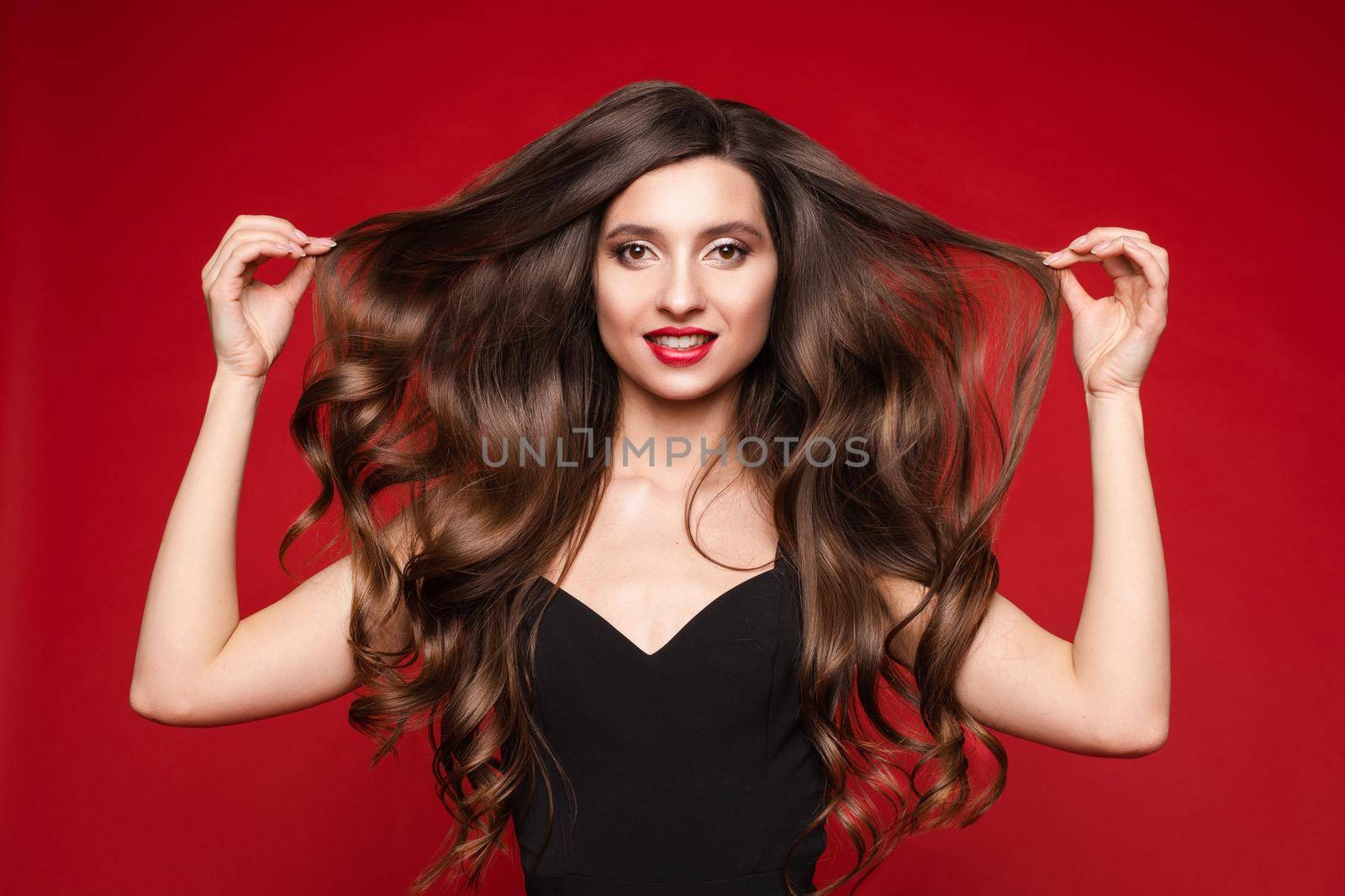 Studio portrait of stunning young caucasian woman with gorgeous long brunette hair in black with red lips. Isolate on red. She is showing beautiful, smooth and healthy hair.