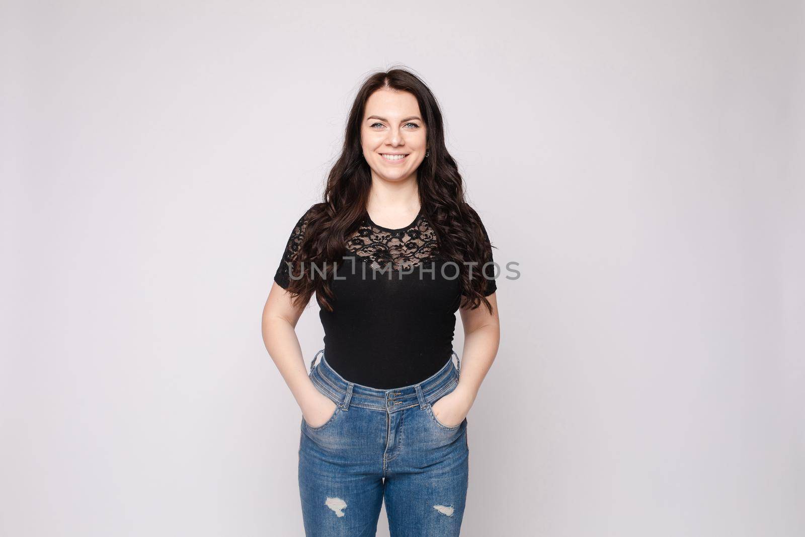 View from front of beautiful slim woman wearing black shirt and jeans standing steady on frey isolated background. Young blonde looking at camera, smiling and posing in studio.