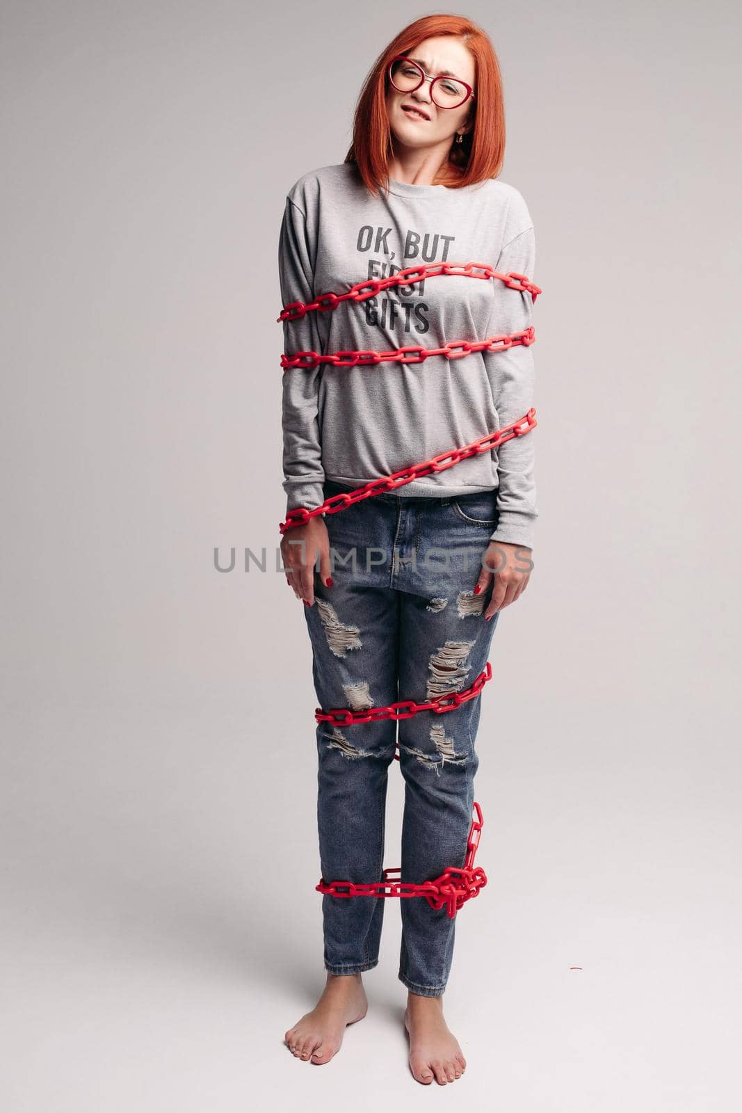 Studio portrait of a girl entangled in a chain. A red-haired woman with brown hair tries to get rid of the chain. The concept of a life-like situation. Isolated on a gray background.