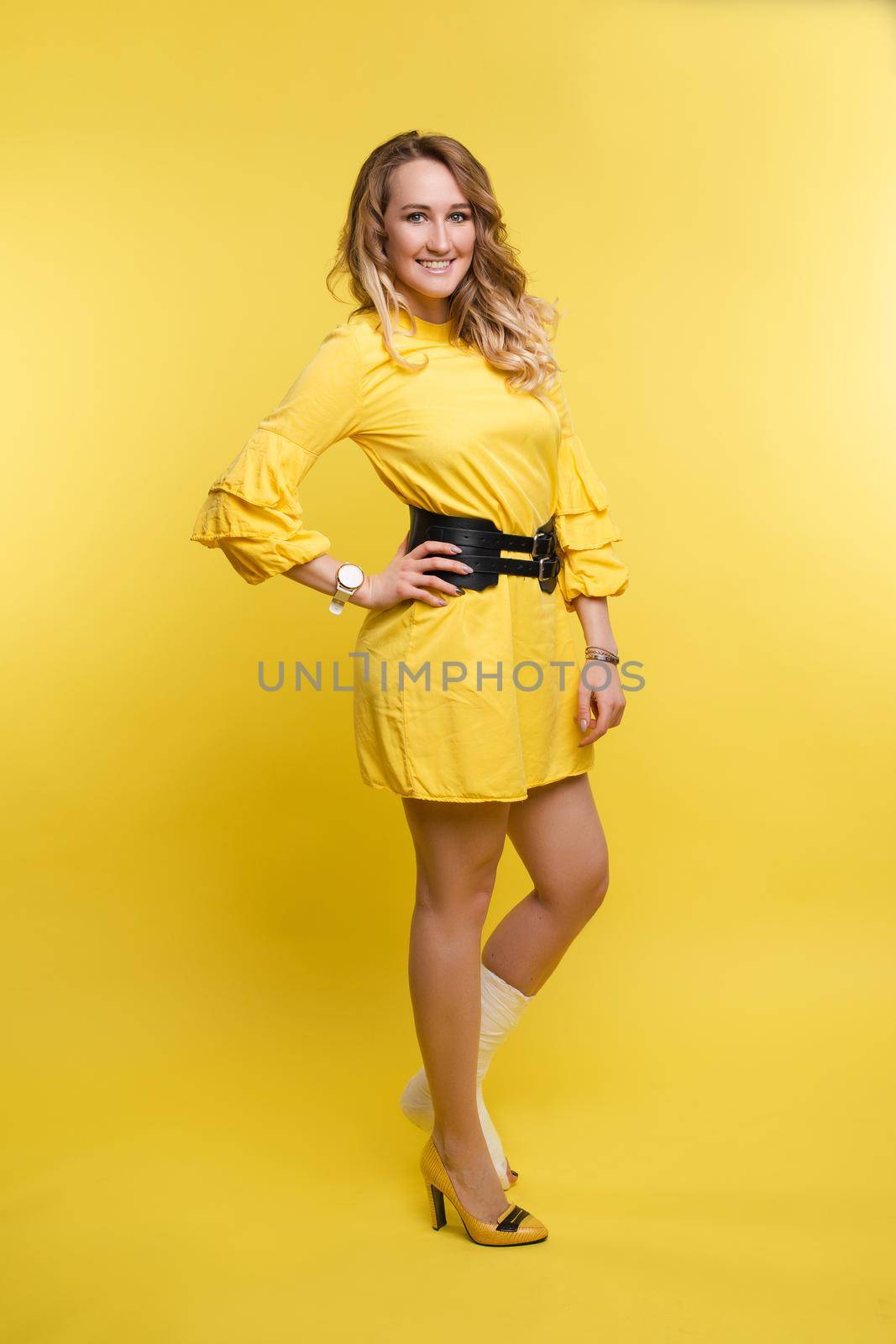 Full length portrait of stunning young woman in yellow dress with one leg in plaster. She is posing with bent broken leg in plaster showing positive attitude. Cheerful lady in dress with plaster. Isolate on red.