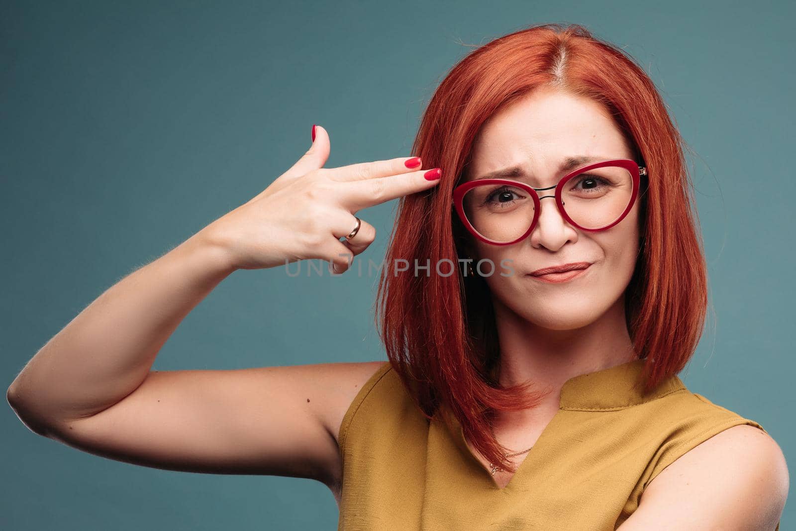 Isolated shot of a young woman head, committing suicide with a finger gesture. Studio portrait of a tired girl shooting herself doing a gun mark with her finger isolated on a blue background. Human facial expressions