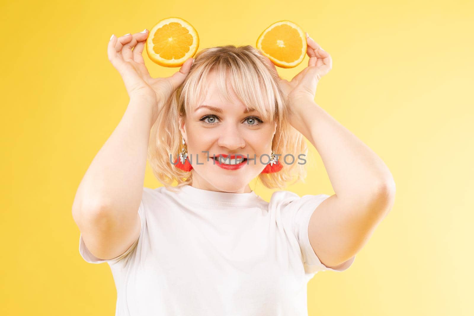 Cropped view of sunny blonde in white shirt looking at camera while posing with fruits on yellow isolated background. Happy girl keeping oranges and smiling. Concept of happiness and summer.