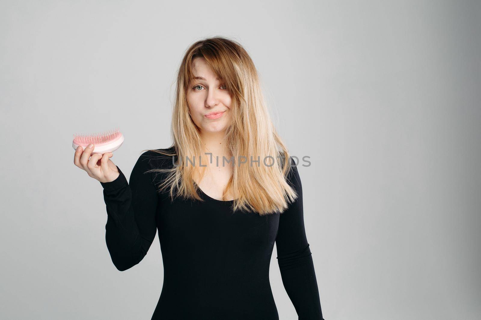 Close up portrait of young confused girl with messy blonde hair. Pretty woman with green eyes, wearing in black sweater, holding hairbrush in hand and looking at camera against white background.