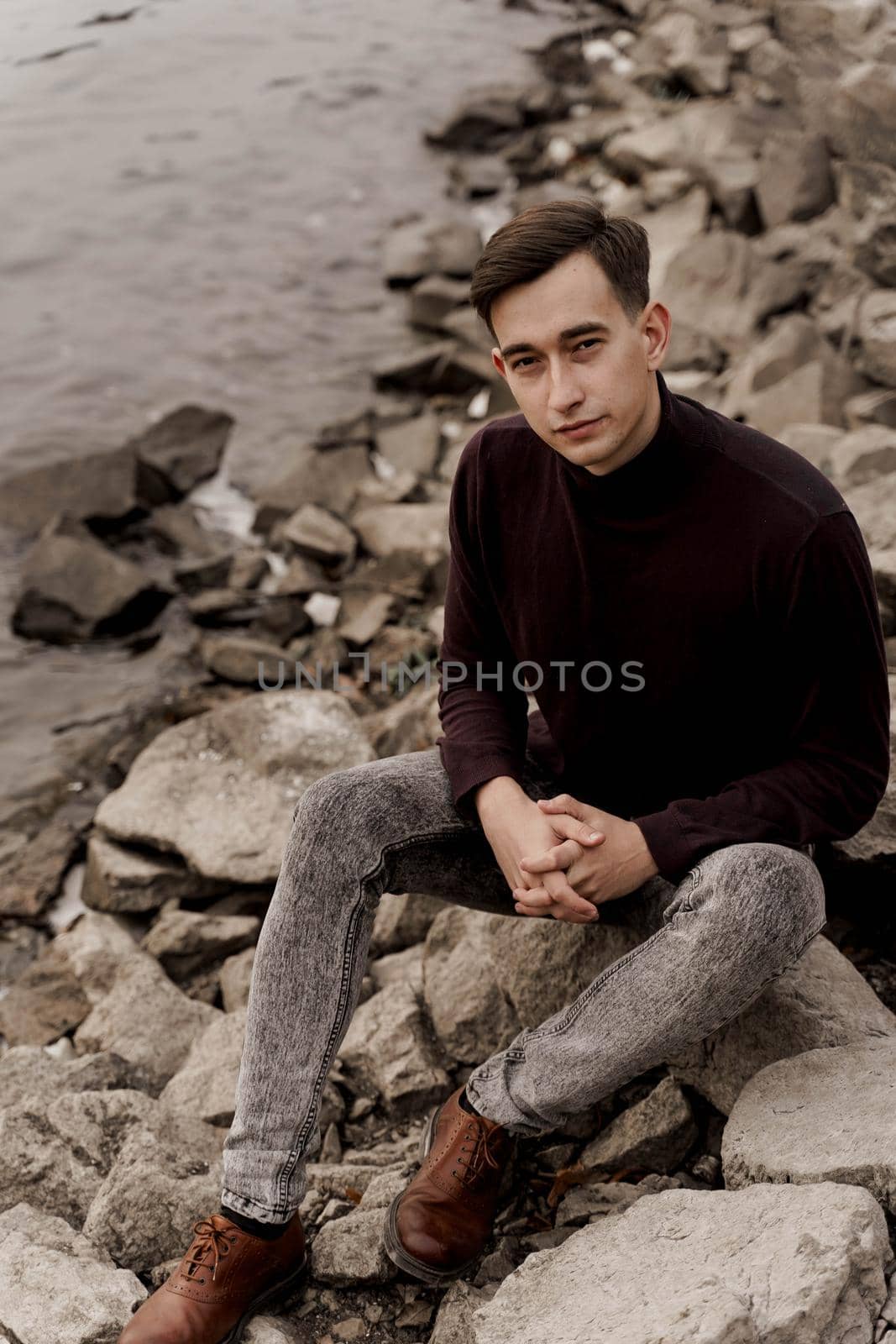 Handsome man seats on the rocks near lake. Professional male model weared formal pullower