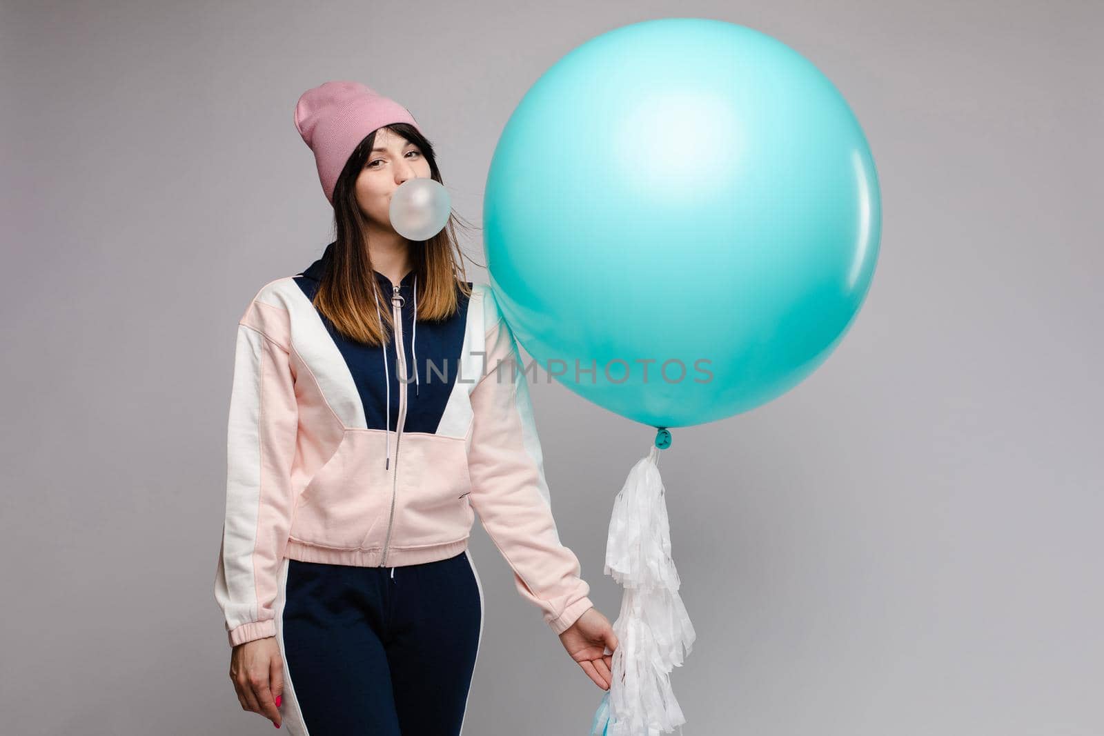 Front view of stylish female teenager wearing tracksuit and hat chewing gum and keeping big blue balloon in studio. Young girl looking at camera and posing on grey isolated background.