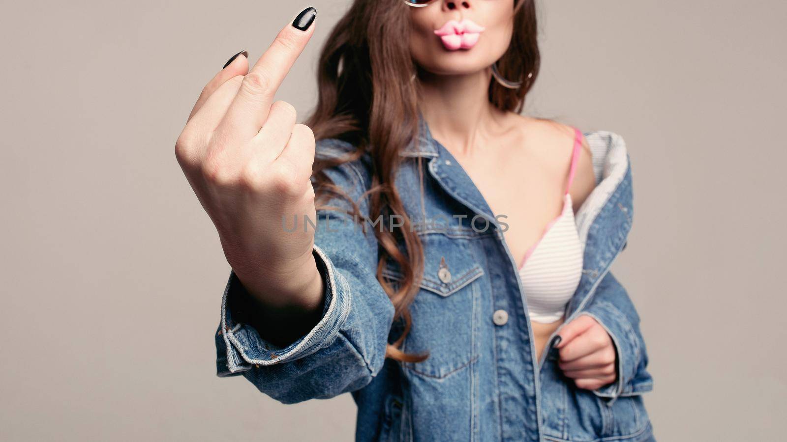 Offensive gesture showing at camera. Stylish young brunette in mirrored sunglasses showing middle finger with black nail. She is wearing denim jacket over a bra. Isolate on grey. Studio shot.