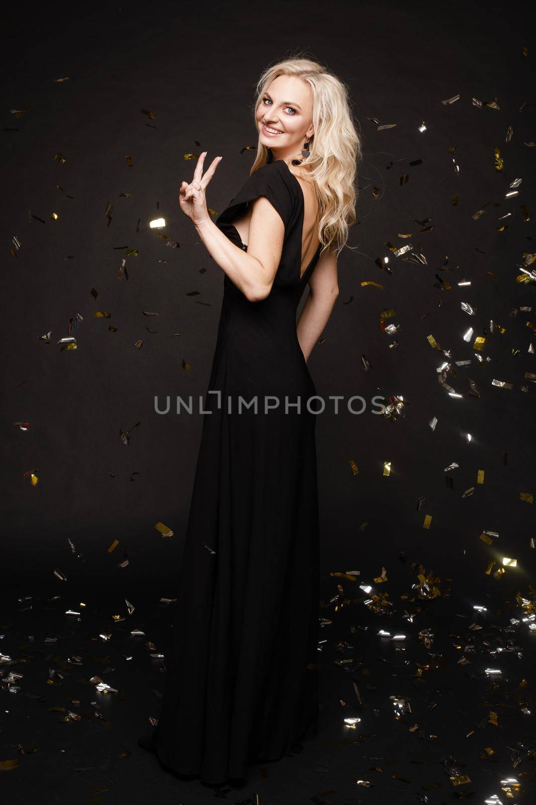 Happy young girl looking surprisingly while golden confetti falling. Beautiful woman in emerald elegant long dress smiling and dancing among decorations. Gorgeous lady celebrating birthday on party.