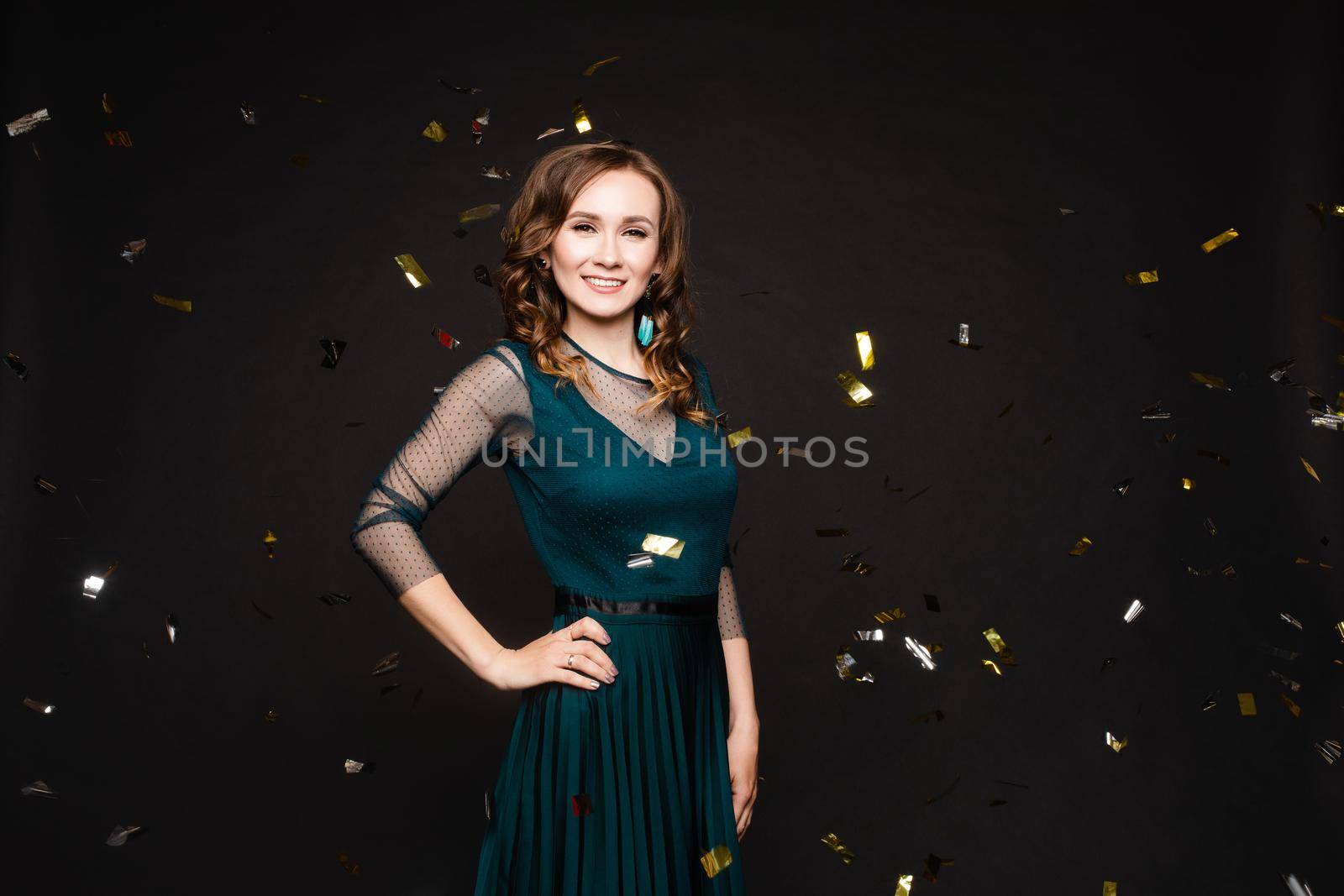 Happy young girl looking surprisingly while golden confetti falling. Beautiful woman in emerald elegant long dress smiling and dancing among decorations. Gorgeous lady celebrating birthday on party.