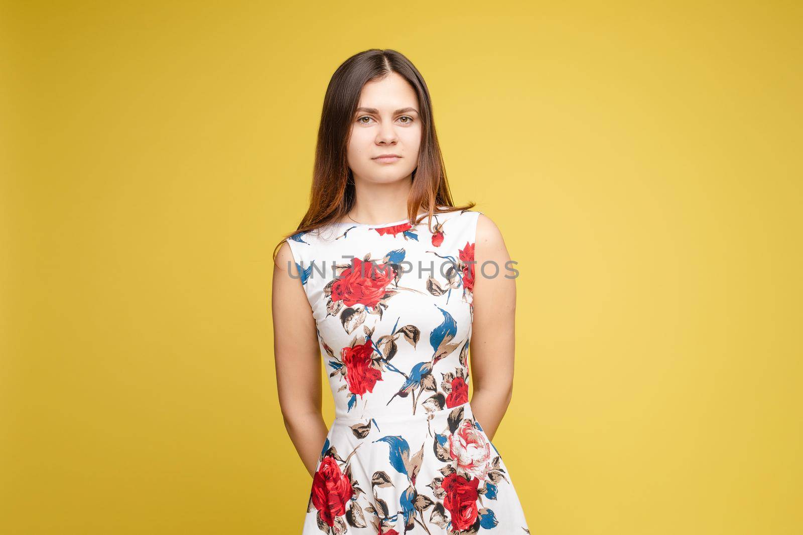 Studio portrait of fashionable brunette lady in white dress with flowers and beige heels posing with bent leg. Smiling at camera. holding skirt. Isolate on yellow background.