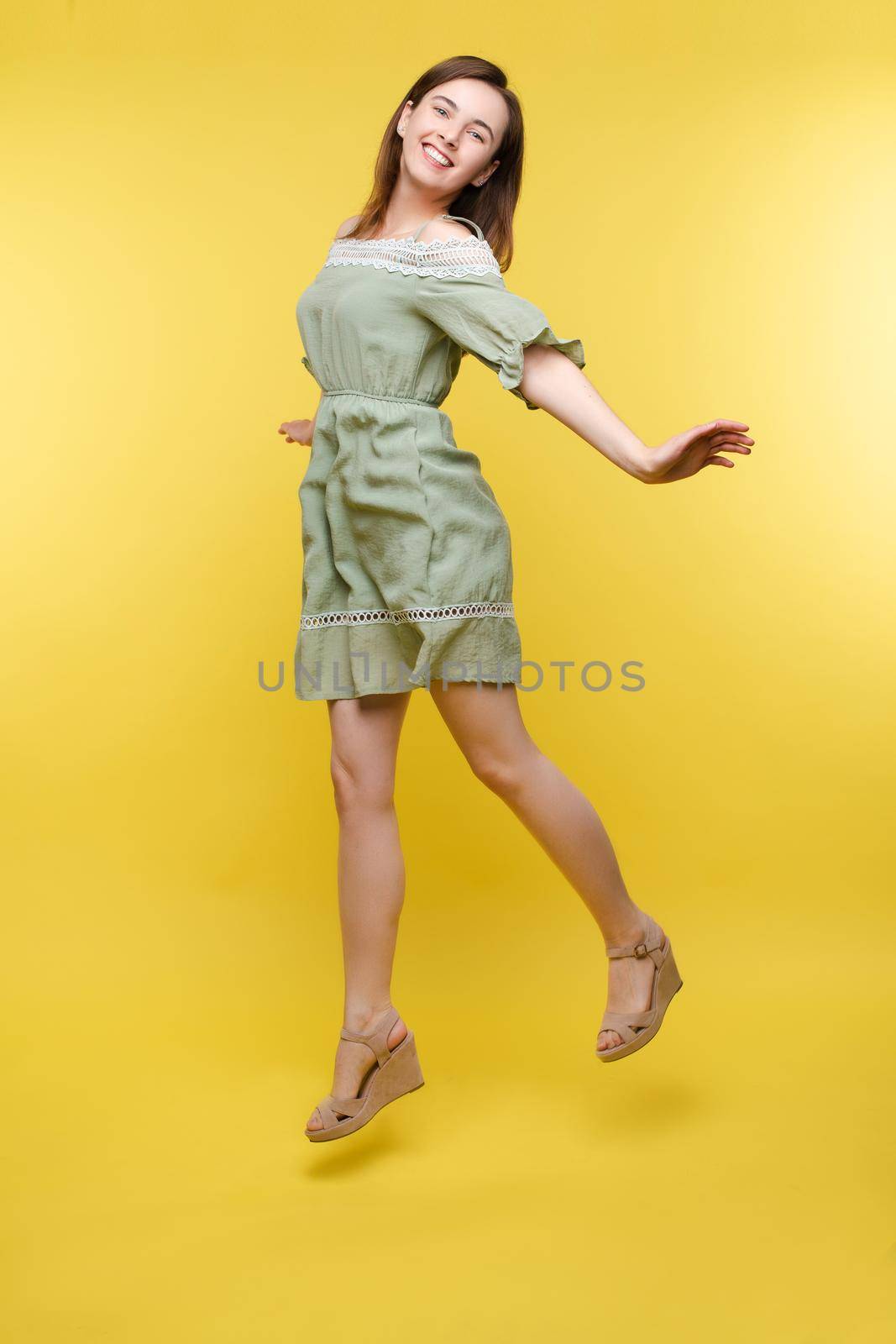 Full length isolate portrait of positive brunette young girl in jeans and t-shirt and sneakers jumping over yellow background. She is smiling at camera.