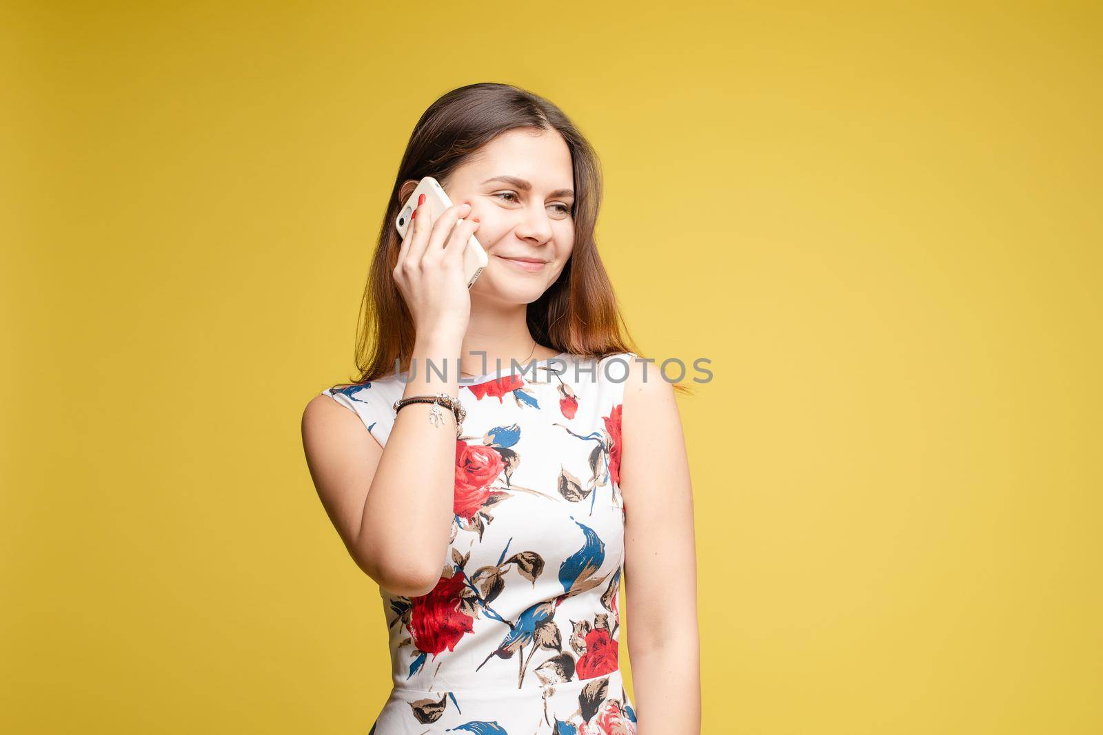 Front view of stylish young girl in bright light dress talking by phone on yellow isolated background in studio. Lovely female chattering and smiling. Concept of communication and happiness.