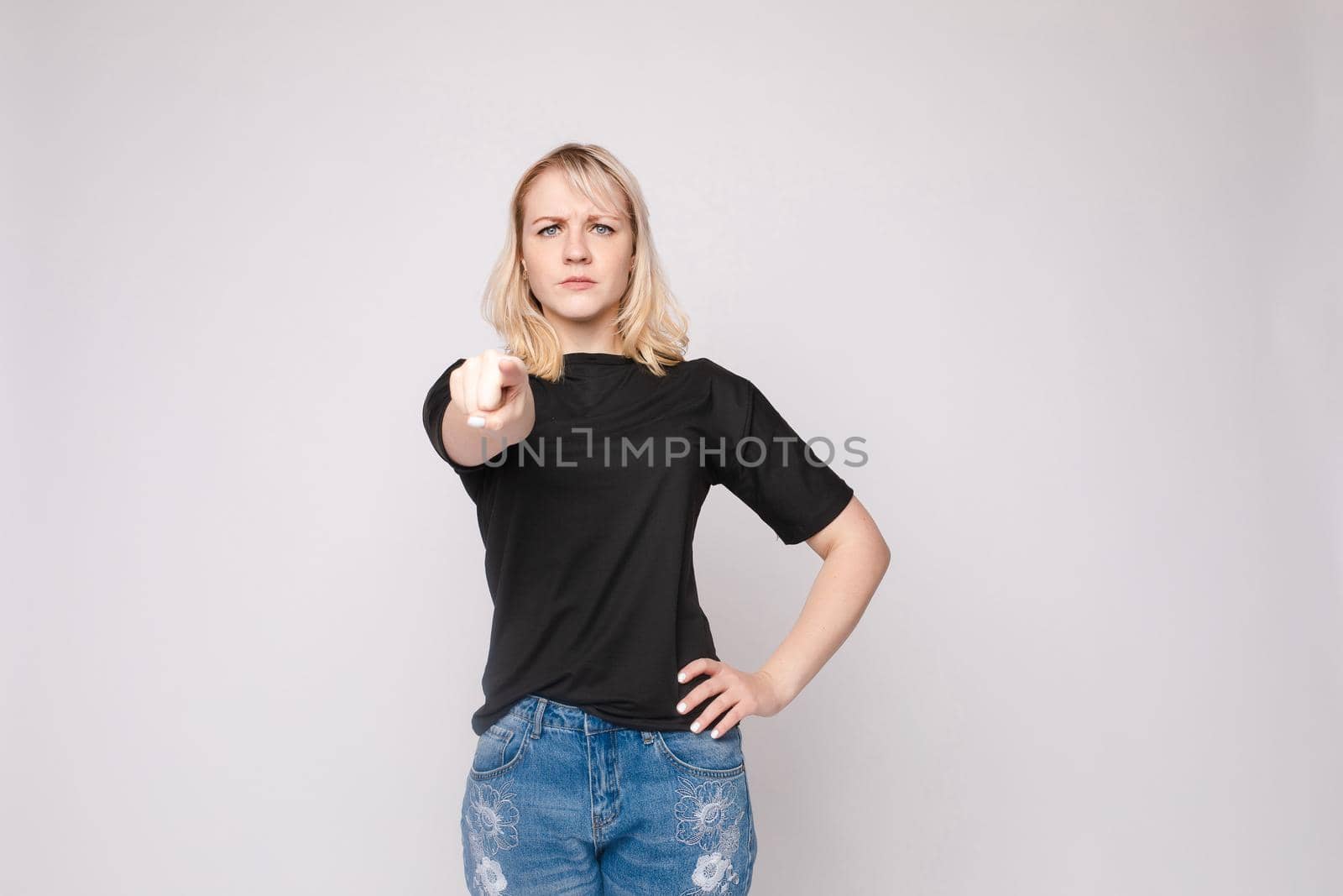 Confident young beautiful woman showing by hand stringent looking at camera posing isolated at white studio background. Accusing female making pointing finger gesture medium long shot