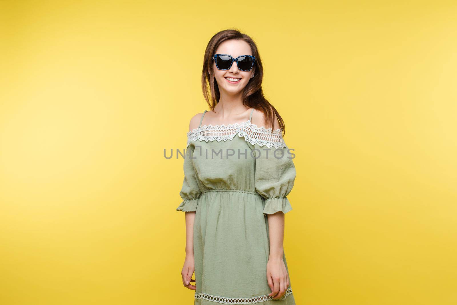 Studio portrait of attractive happy and jovial Caucasian brunette woman in plain pistachio dress and trendy black sunglasses smiling at camera against yellow background.