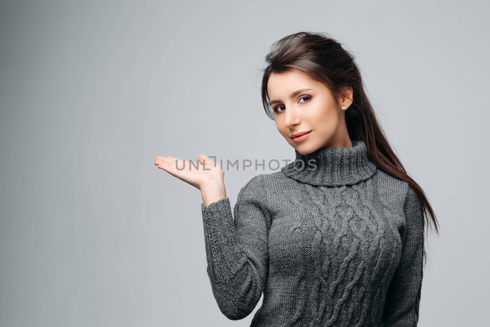 Waist up portrait of beautiful girl showing empty copy space in left side. Pretty brunette with stylish hairdo and perfect makeup. Isolated on gray background