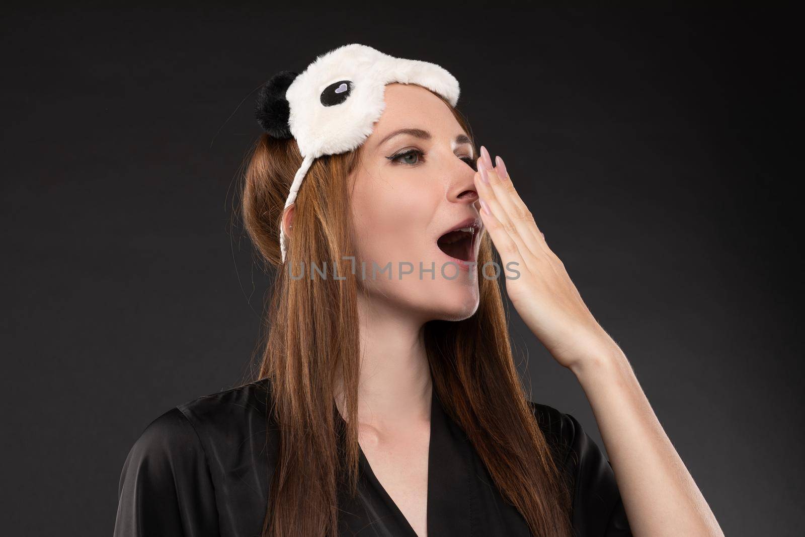 Young attractive girl in black elegant pajamas going to bed after hard working day. Beautiful woman with eyes mask on head ready for relaxing. Brunette pretty lady waking up and yawning in morning.