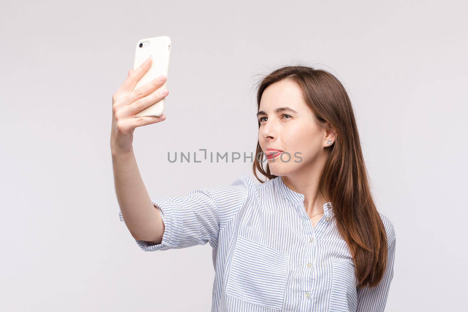 Studio portrait of pretty young Caucasian girl with long brunette hair in casual striped shirt making funny face and sticking tongue at camera while making selfie on cell phone..