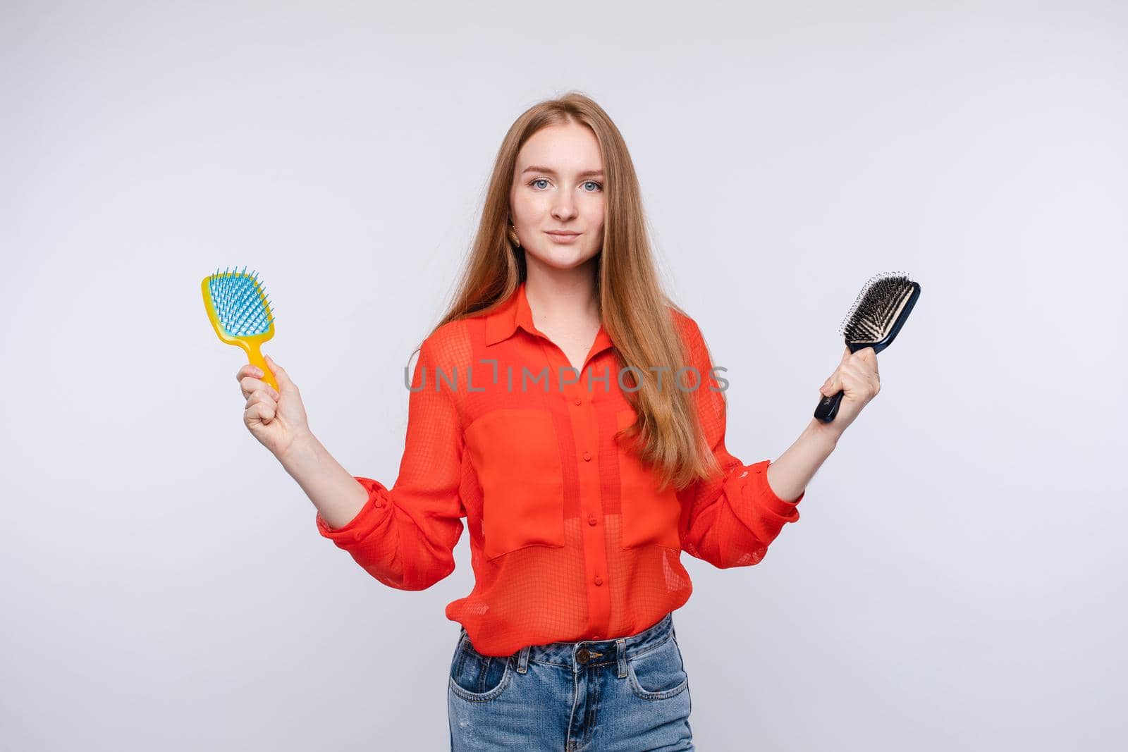 Front view of young woman keeping in hands two brushes and choosing hair care. Longhaired girl looking at camera and thinking on grey isolated background. Concept of choice and copy space.