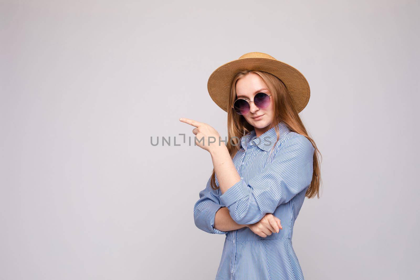 Studio portrait of stylish woman with redhair in sunglasses, hat and checked shirt dress indicating blank space in the air with her index finger.