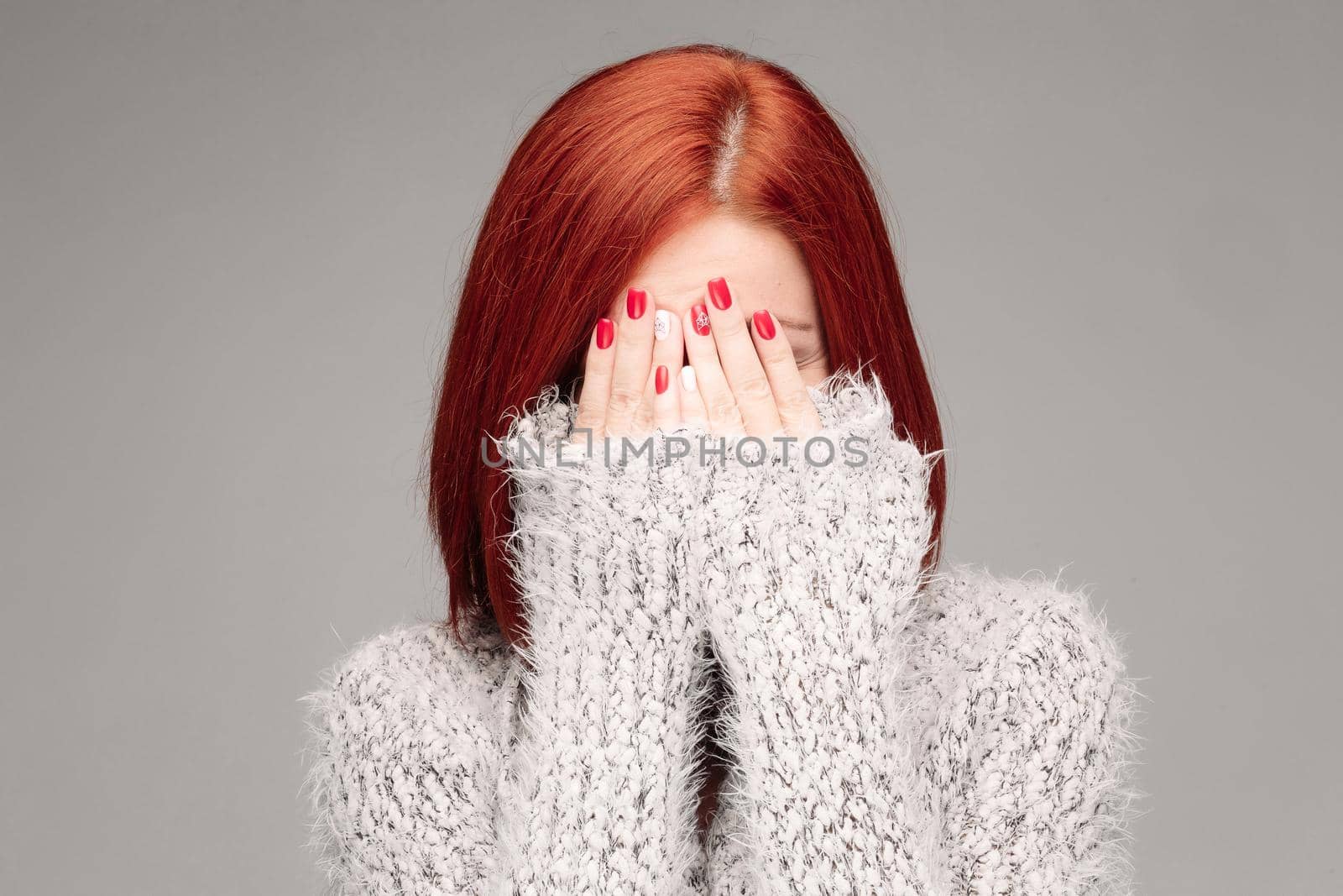 Studio portrait of young red haired unrecognizable woman wearing warm gray seater hiding face by hands. Incognito girl having reaction of protection or saving secret. Gray studio background.