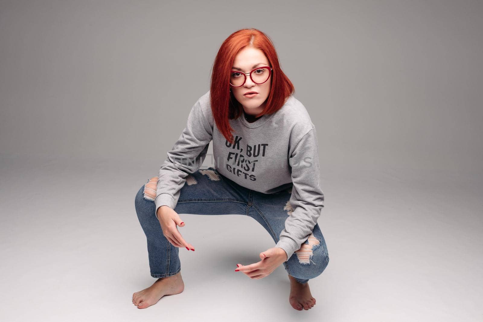 Swag and confident red haired girl in jeans and sweatshirt posing at gray studio, looking at camera and gesturing by hands. Young stylish woman wearing in bag girl look. Concept of fashion.