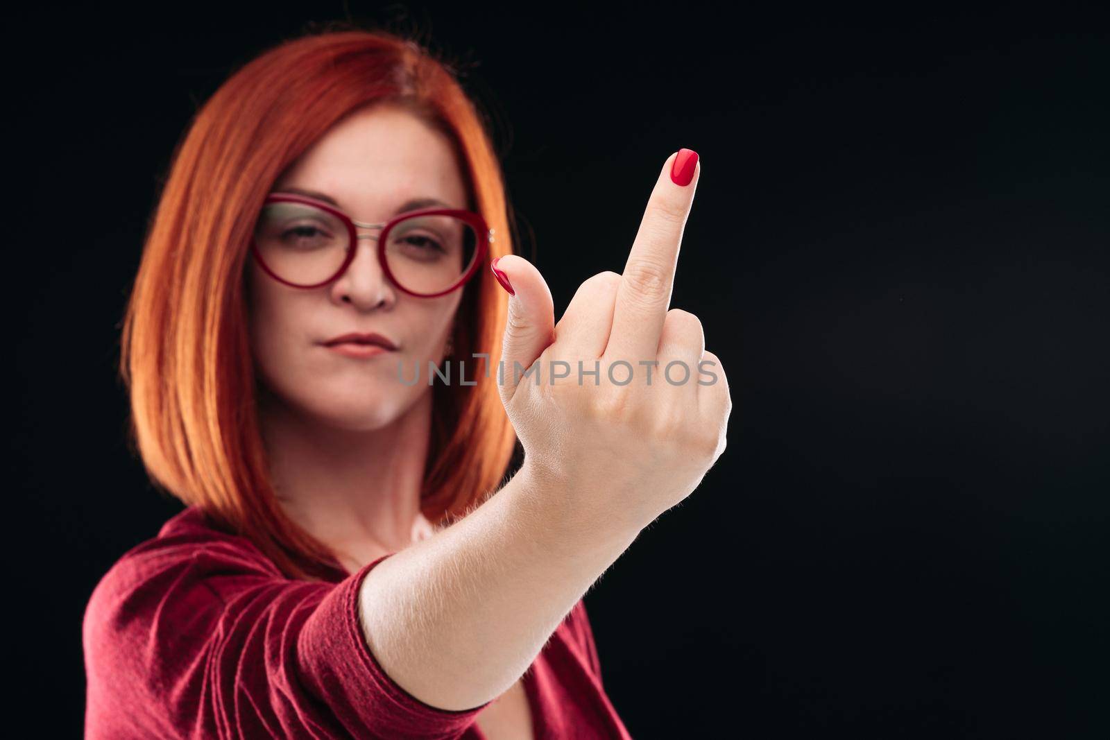 Confident and angry red haired girl with short hairstyle wearing glasses showing finger in camera. Emotionally young woman posing, gesturing fuck off, out by hand. Black studio background.