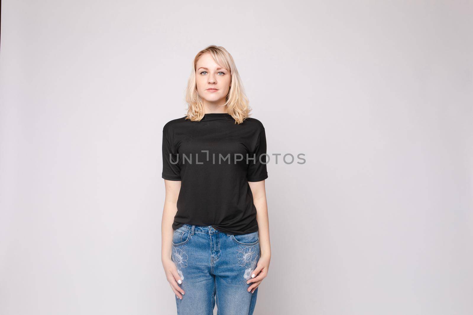 View from front of beautiful slim woman wearing black shirt and jeans standing steady on frey isolated background. Young blonde looking at camera, smiling and posing in studio.