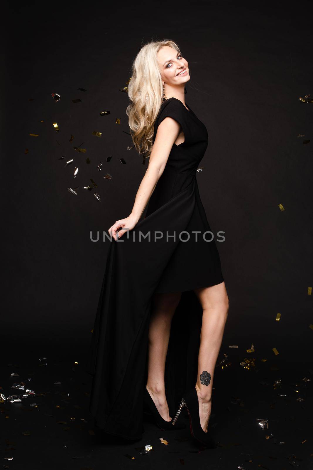 Front view of elegant woman wearing long black dress looking at camera and smiling on black isolated background. Attractive blonde with long legs posing in studio. Concept of party and beauty.