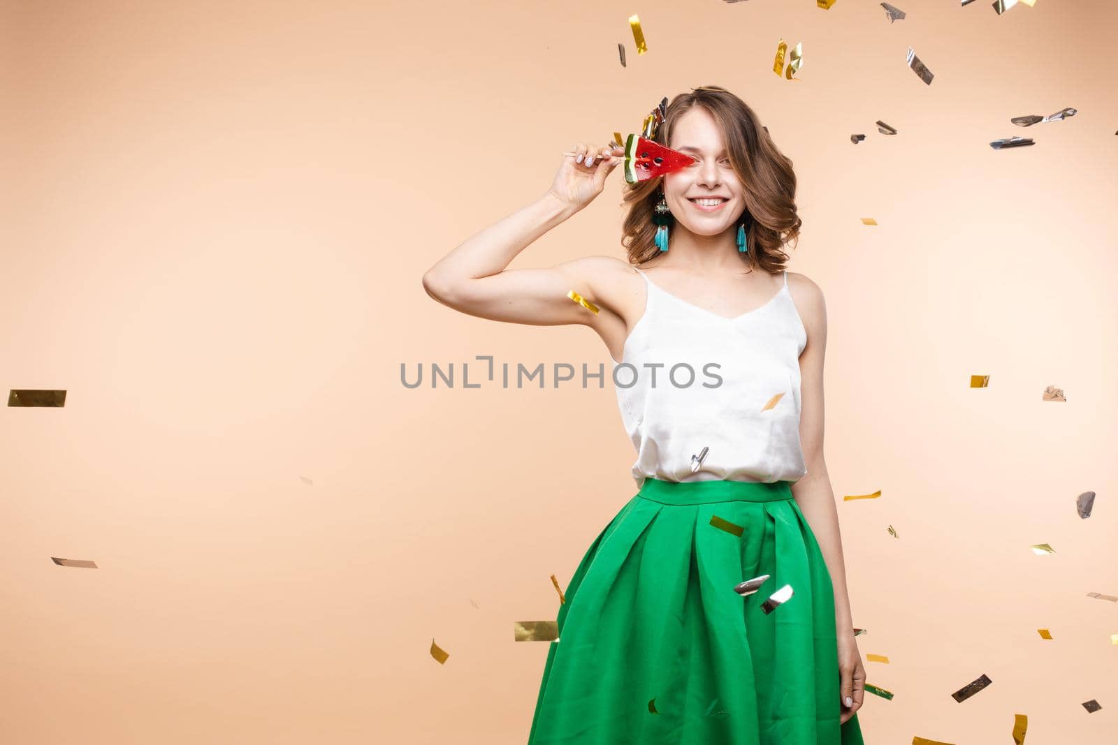 Playful elegant smiling woman posing with piece watermelon candy surrounded shining colorful confetti. Happy young female having positive emotion looking at camera isolated at light studio background