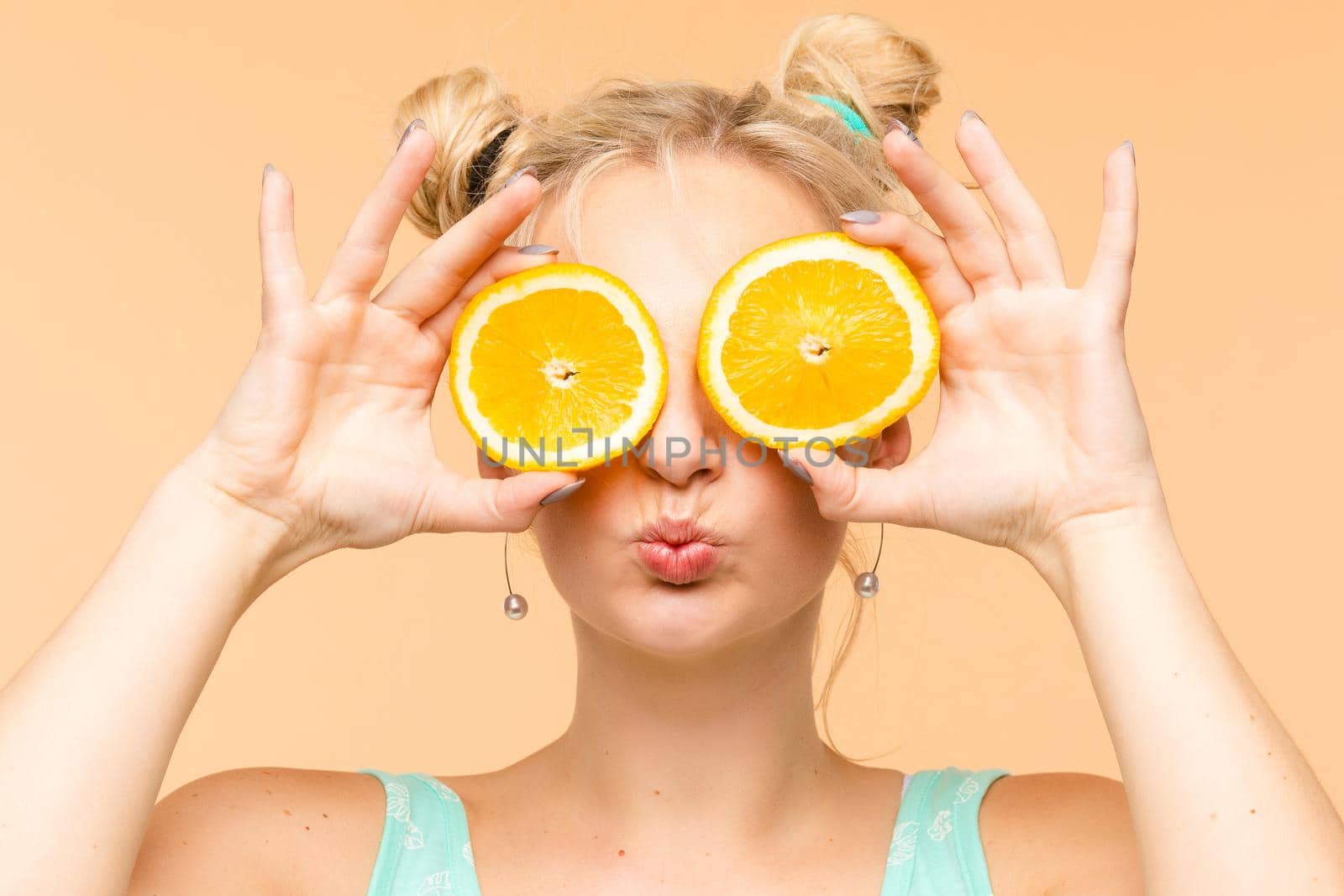 Front view of cheerful young blonde posing with fresh oranges on isolated background. Funny girl keeping fruit and closing eyes in studio. Concept of happiness and health.