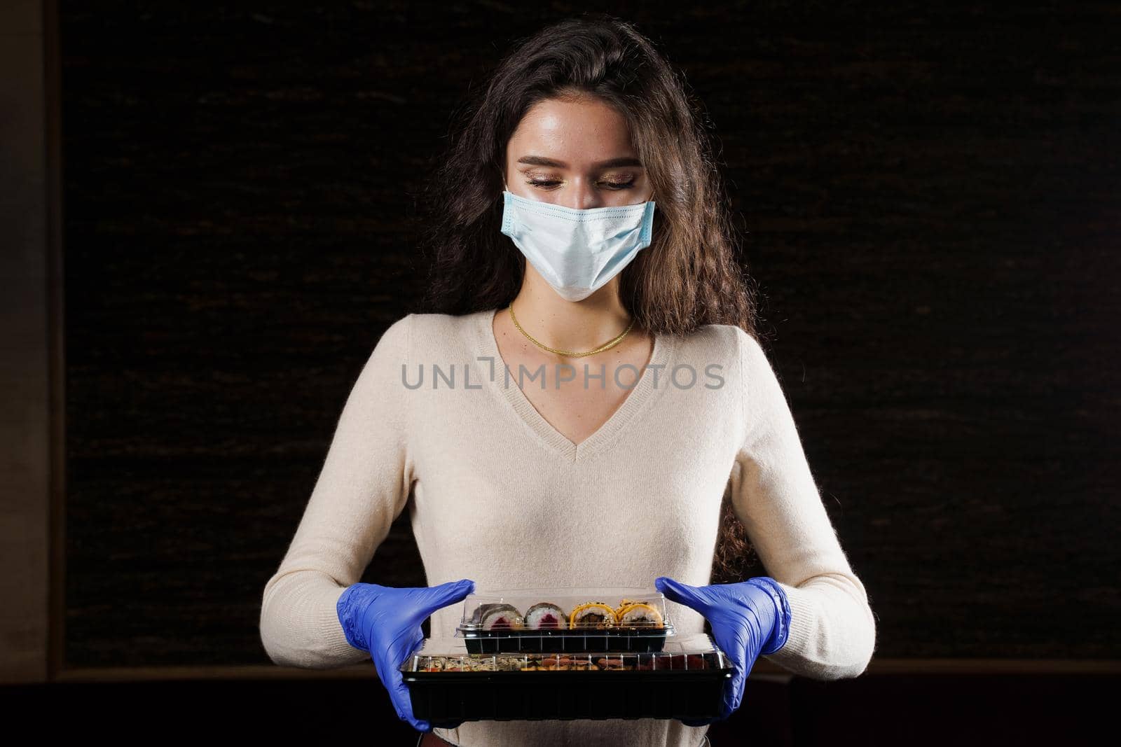 Girl with sushi. Food courier in medical mask. Delivery at quarantine coronavirus covid-19. Young woman holding 2 sushi boxes in hands using gloves. by Rabizo