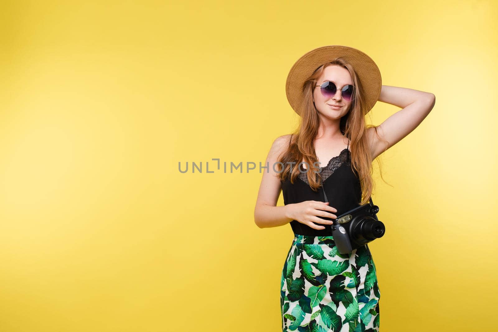 Front view of bright young girl wearing straw hat and glasses posing on yellow isolated background in studio. Female tourist keeping camera, looking at camera and smiling. Concept of summer.