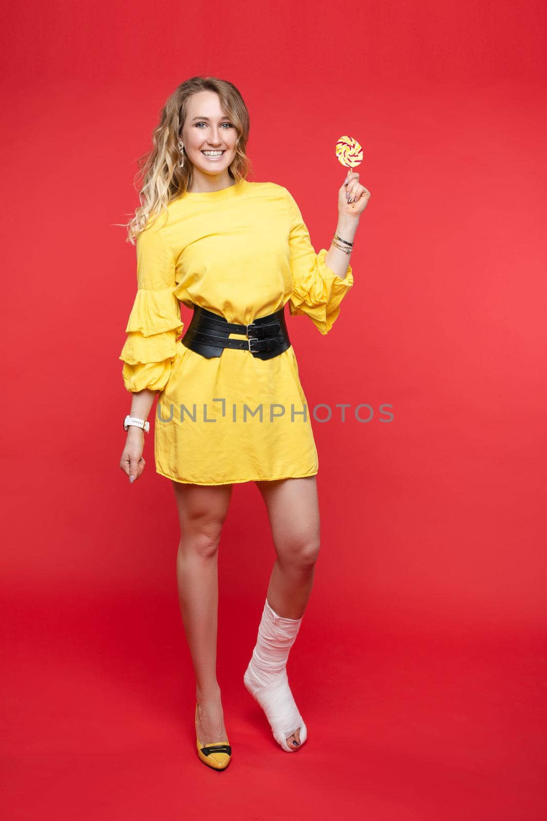 Studio portrait of confused brunette in fashionable summer yellow dress with belt looking at sweet lollipop with suspicion. Isolate on red background. She wants sweet but cannot eat it.