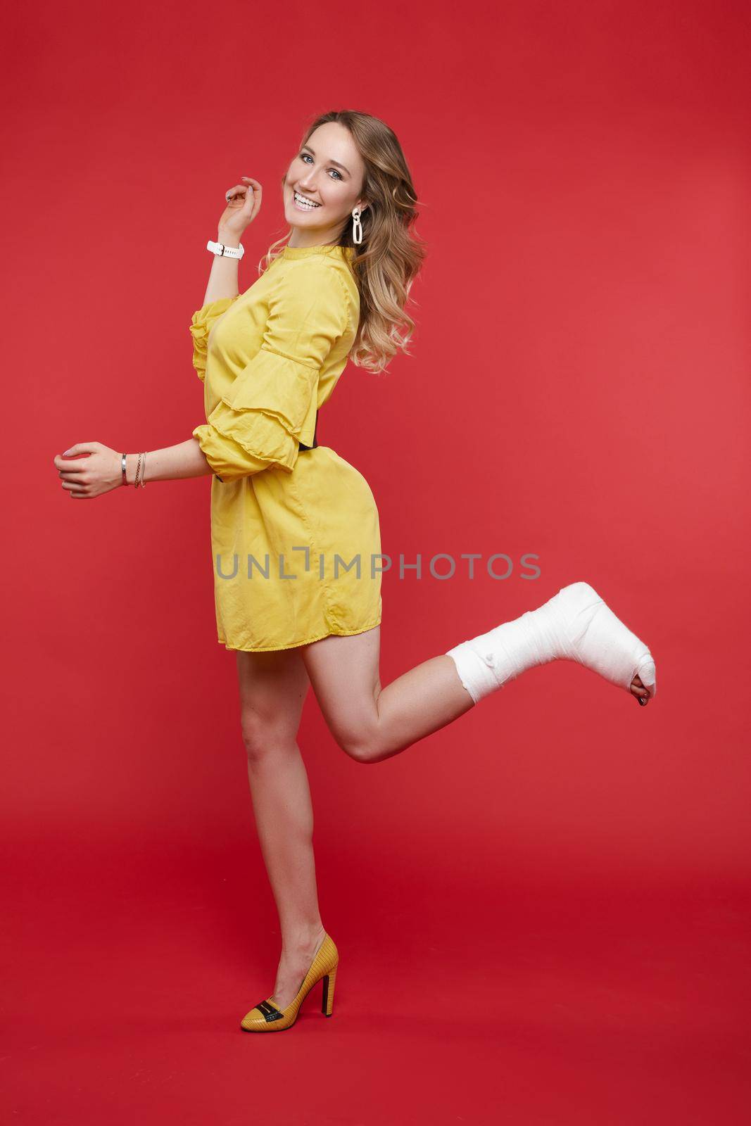 Full length portrait of stunning young woman in yellow dress with one leg in plaster. She is posing with bent broken leg in plaster showing positive attitude. Cheerful lady in dress with plaster. Isolate on red.