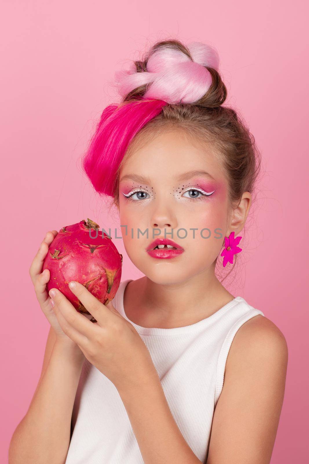Close-up portrait of pretty girl with pink hairstyle with dragon fruit on pink background. Studio shot of charming tween girl with pink make up enjoying juicy red pitaya. exotic Pitahaya fruit.