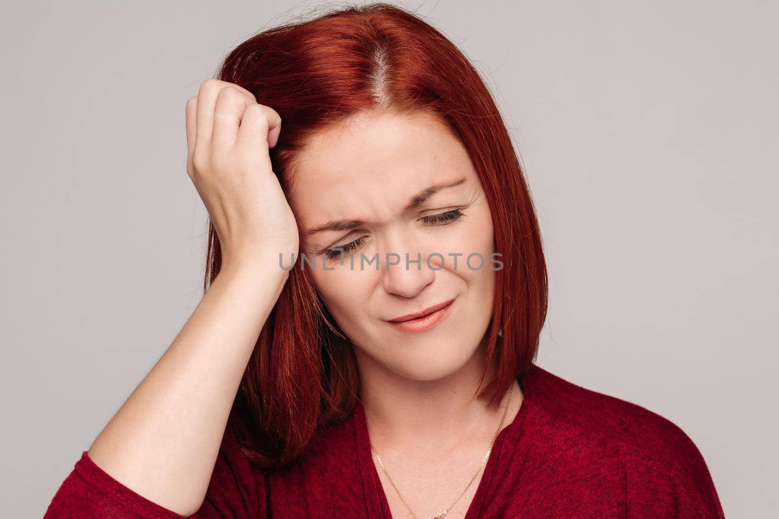 Beautiful young lady with closed eyes holding her hand near head. Clouse up of pretty red haired woman in bordo blouse with headache. Sadly girl touching her hair by hand and thinking about problem.