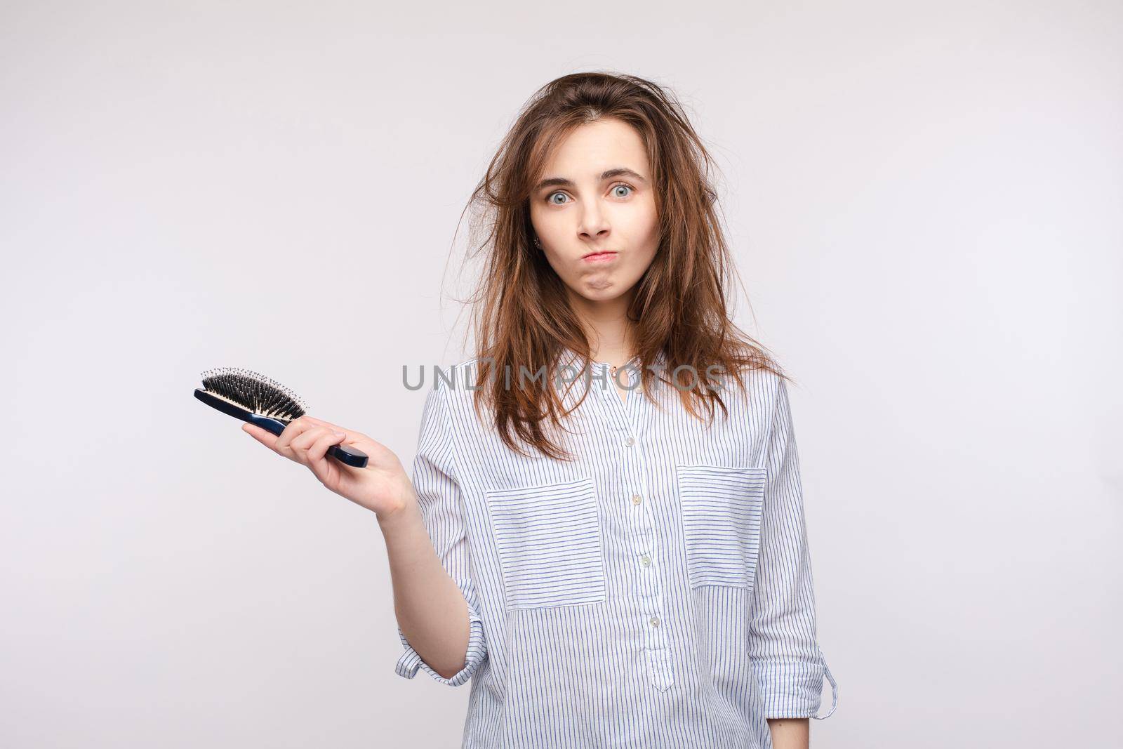 Studio portrait of young adult woman with messy brunette hair holding a hairbrush in right hand and looking at camera with puzzlement and confusion. Isolate on white.