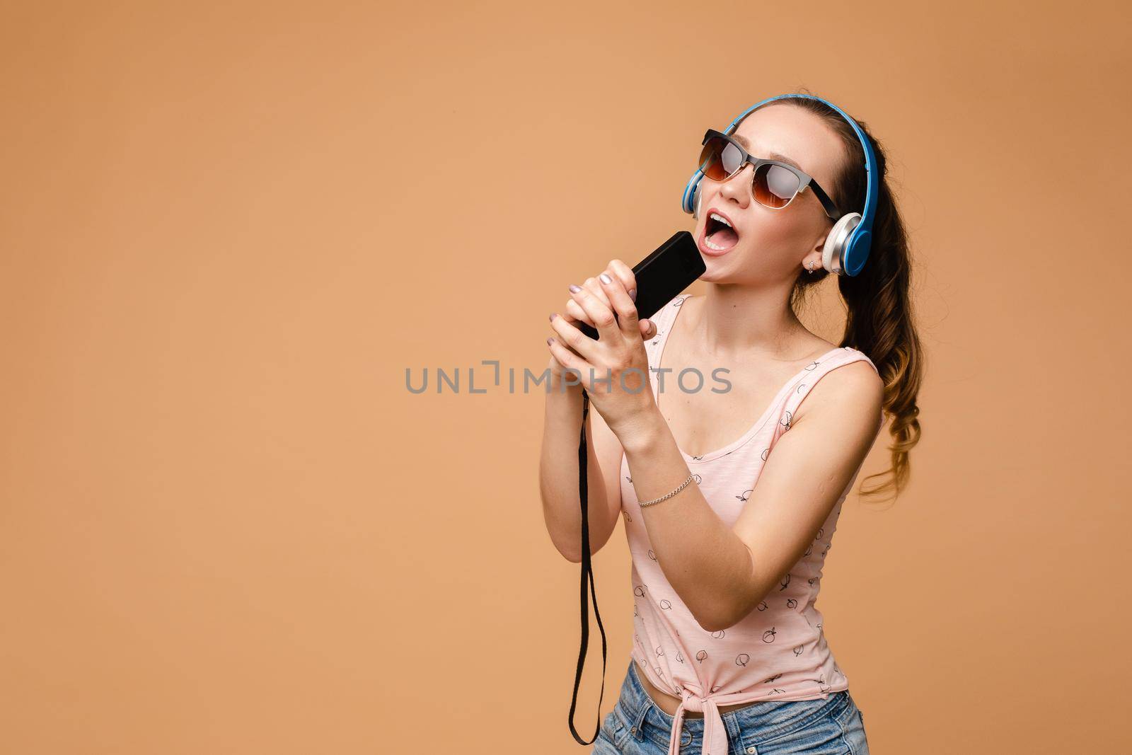 Happy young woman wearing headphones listening music and dancing on isolated background. Cheerful girl in glasses keeping microphone and singing in studio. Concept of relax and fun.