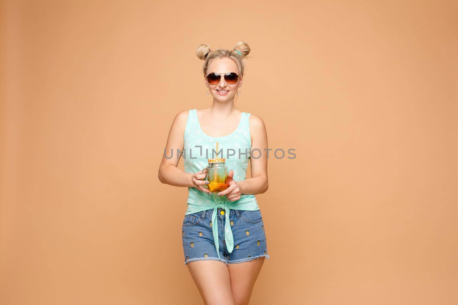 Young happy lady standing on one leg and holding other one up. Blond girl in casual clothes enjoying tasty fruit cocktail. Beautiful woman in sunglasses holding jar with citrus lemonade in hands.