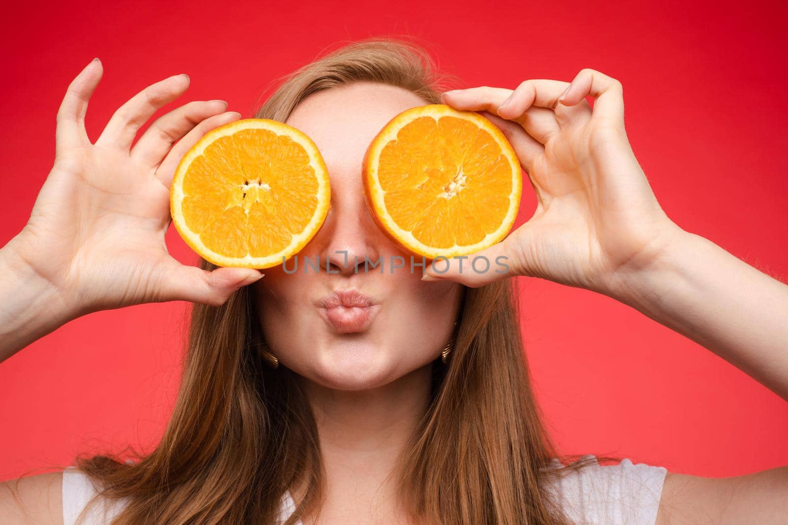 Front view of cheerful young blonde posing with fresh oranges on isolated background. Funny girl keeping fruit and closing eyes in studio. Concept of happiness and health.