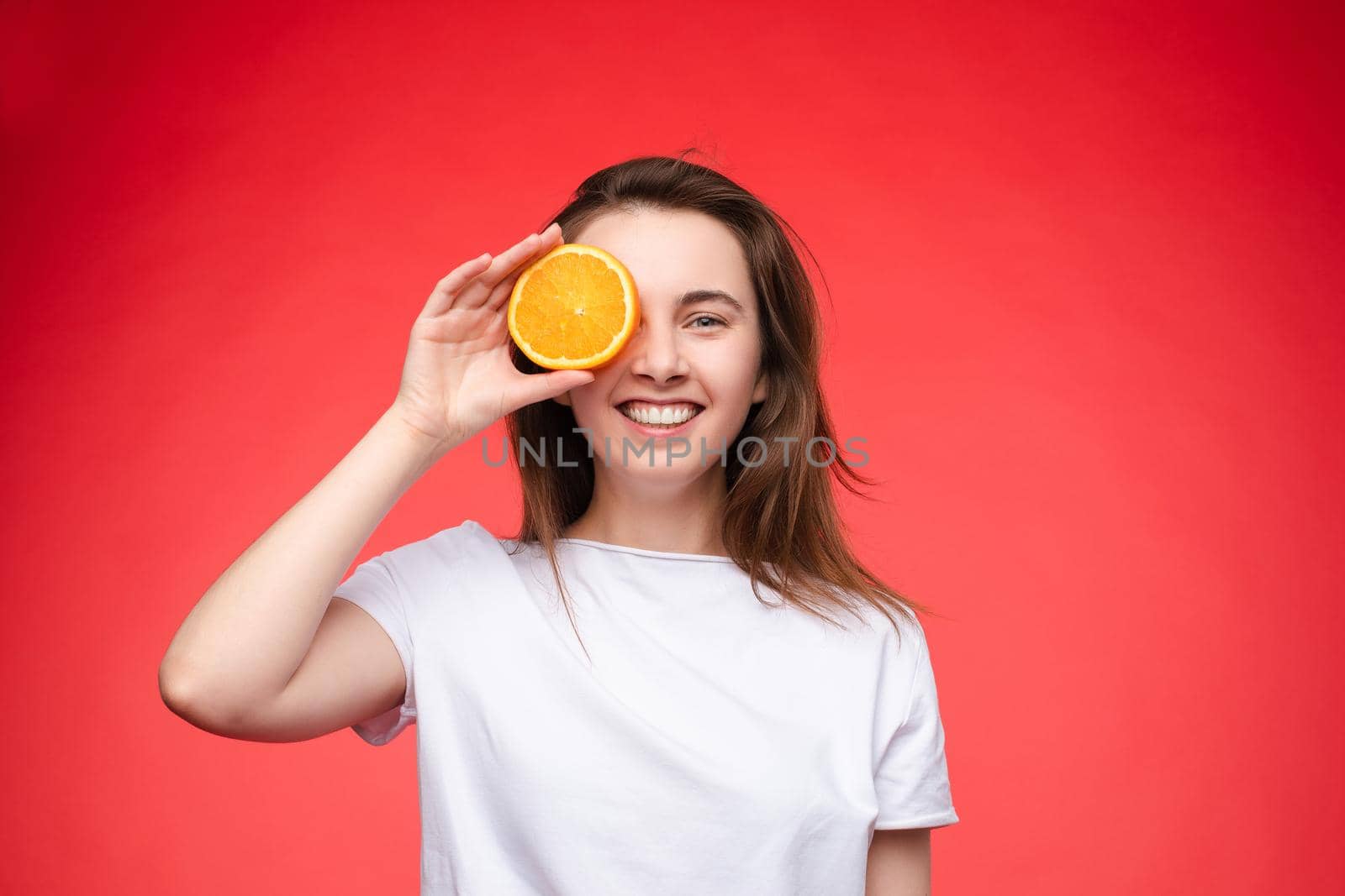 Front view of cheerful young blonde posing with fresh oranges on isolated background. Funny girl keeping fruit and closing eyes in studio. Concept of happiness and health.