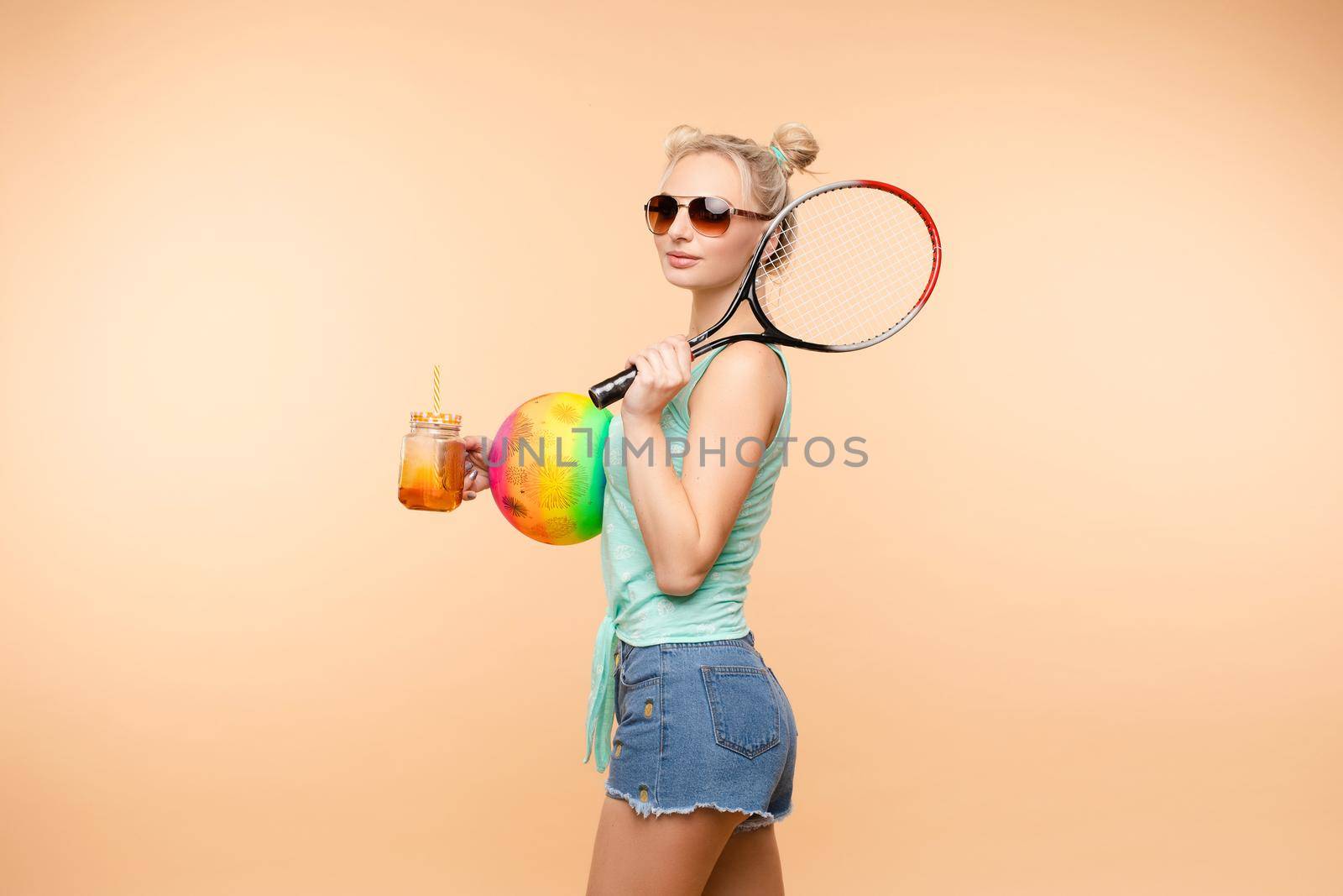 Side view of sporty blonde drinking fresh juice, doing sport and posing in studio. Young model keeping ball and racket and smiling on isolated background. Concept of energy and power.