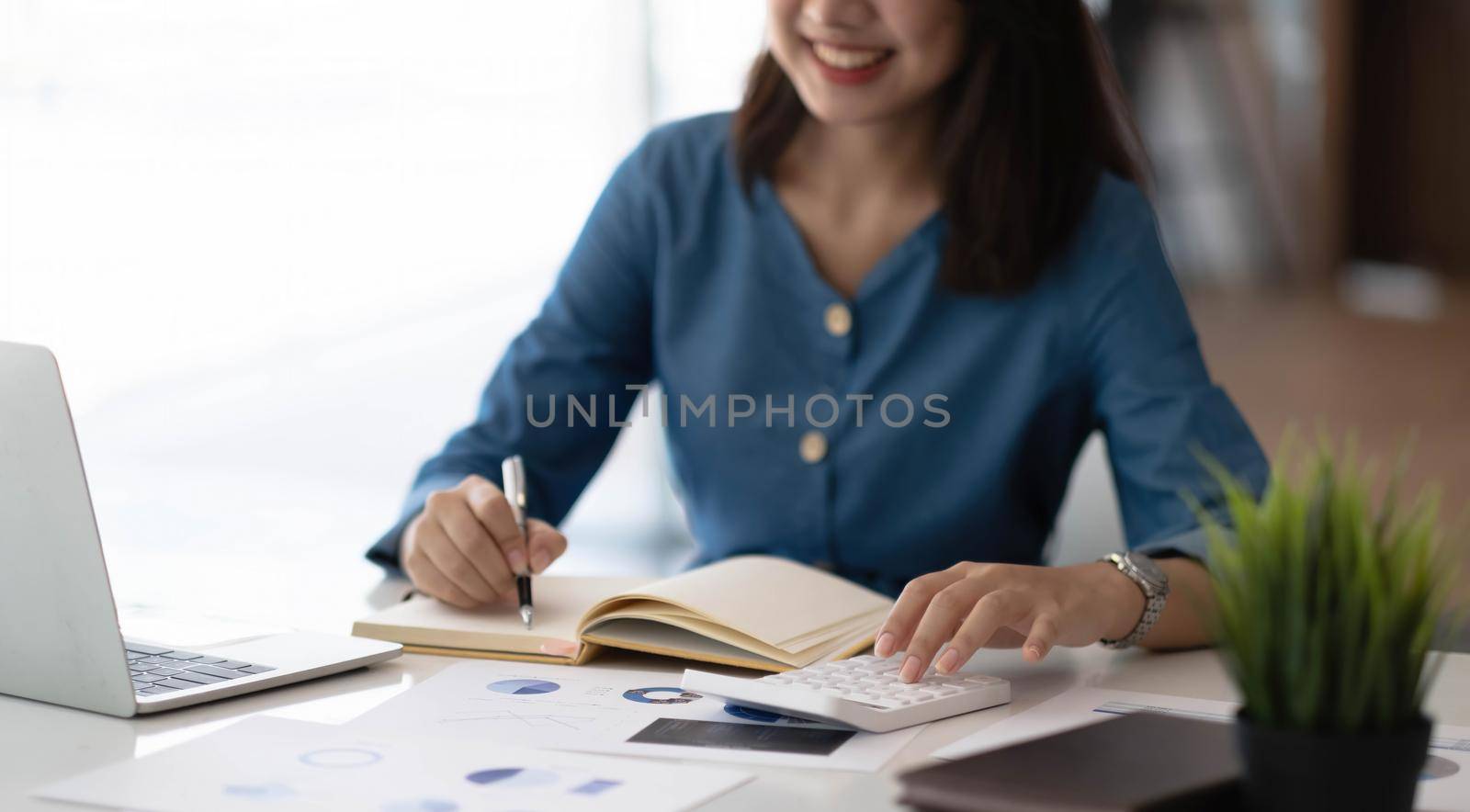 Cheerful young woman with laptop and jar and writing in notebook while sitting at table and working on freelance project at home by wichayada