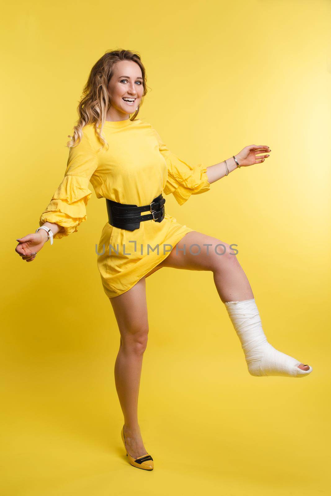 Full length portrait of stunning young woman in yellow dress with one leg in plaster. She is posing with bent broken leg in plaster showing positive attitude. Cheerful lady in dress with plaster. Isolate on red.
