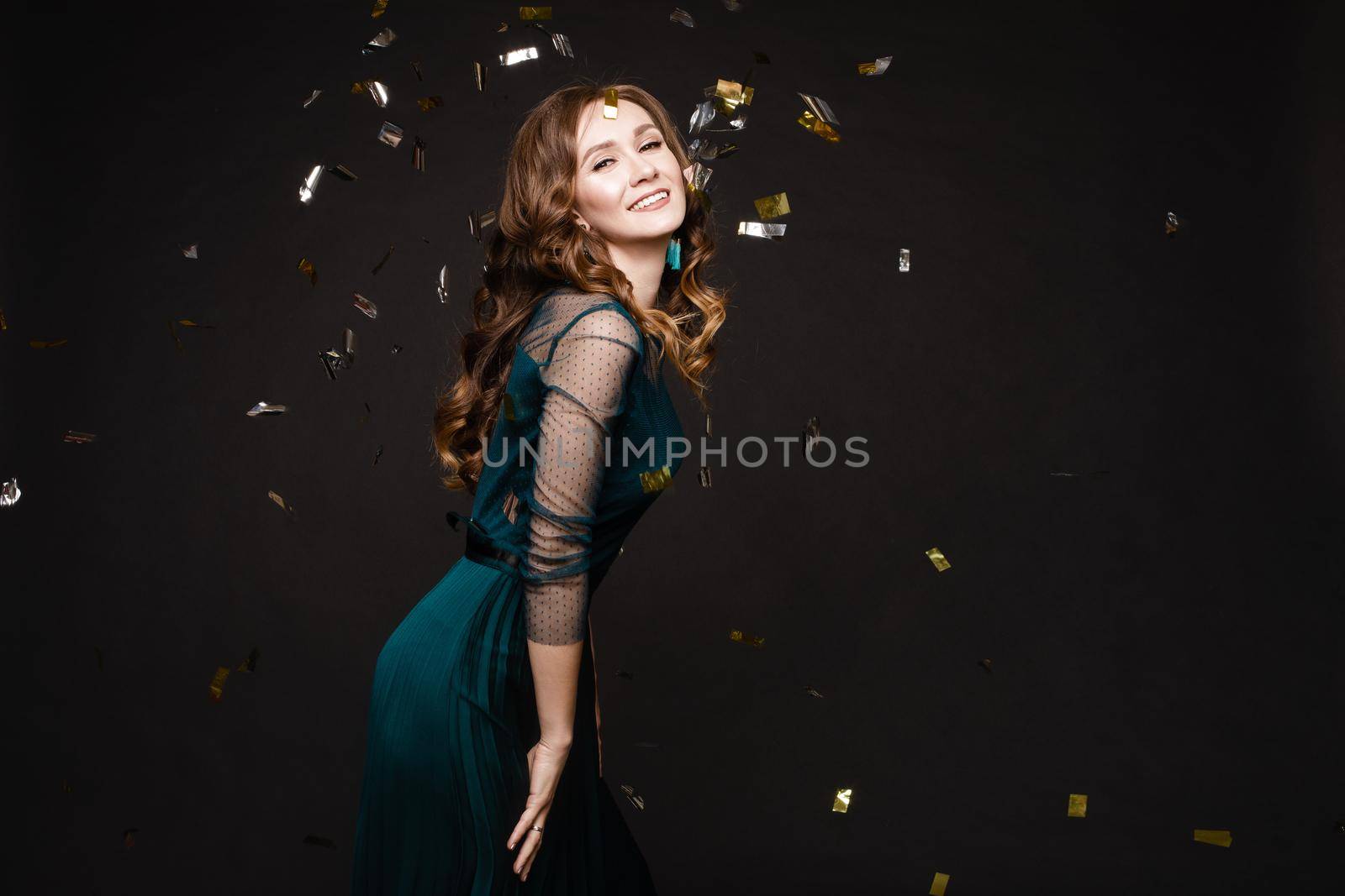 Happy young girl looking surprisingly while golden confetti falling. Beautiful woman in emerald elegant long dress smiling and dancing among decorations. Gorgeous lady celebrating birthday on party.