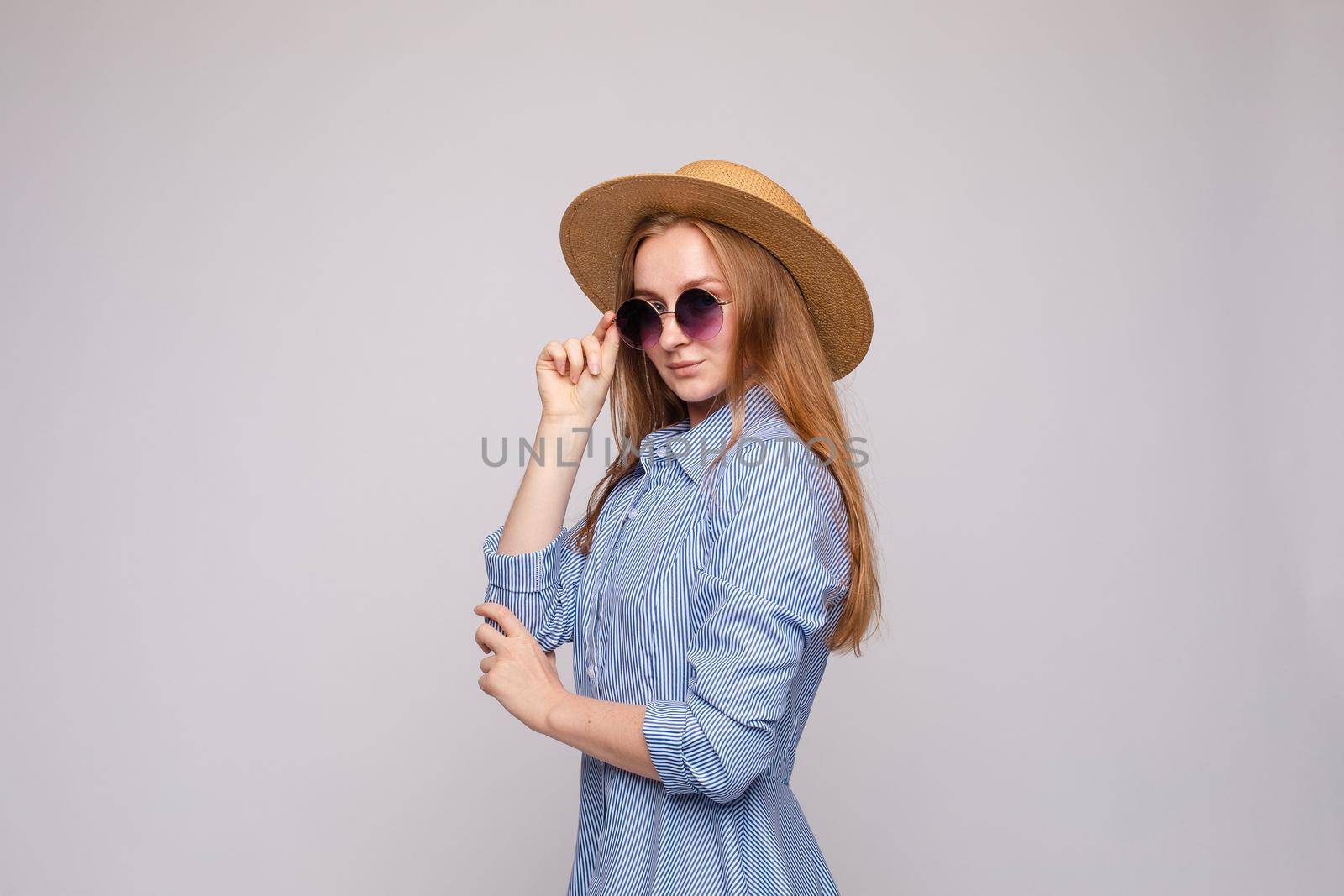 Side view of stylish female model wearing dress, glasses and straw hat posing on grey isolated background. Young blonde looking at camera while standing in studio. Concept of summer outfit.