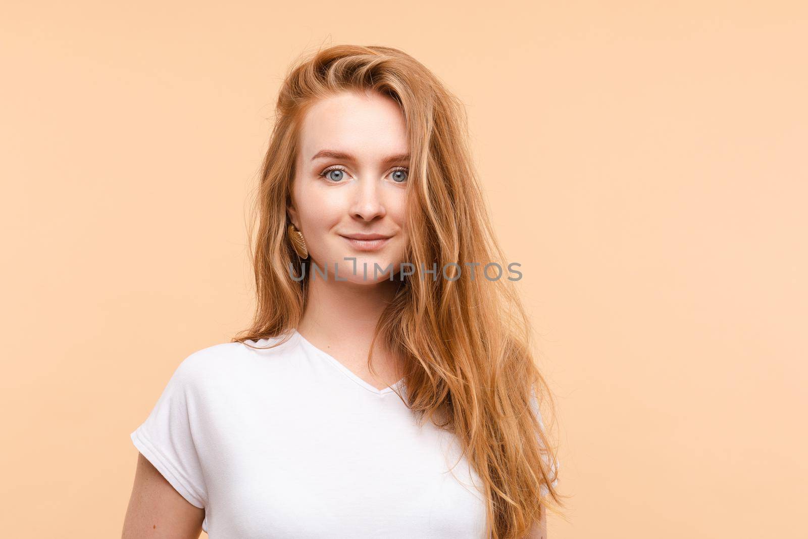 Studio portrait of lovely beautiful young adult woman with blue eyes and long wavy red hair in white t-shirt looking at camera. Isolate on pale yellow background.