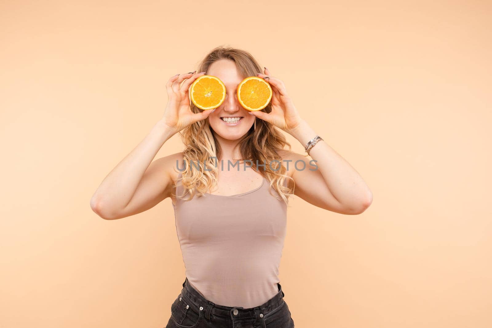 Studio headshot of young funny brunette with hairstyle and red lips holding halved oranges on eyes against bright yellow background.